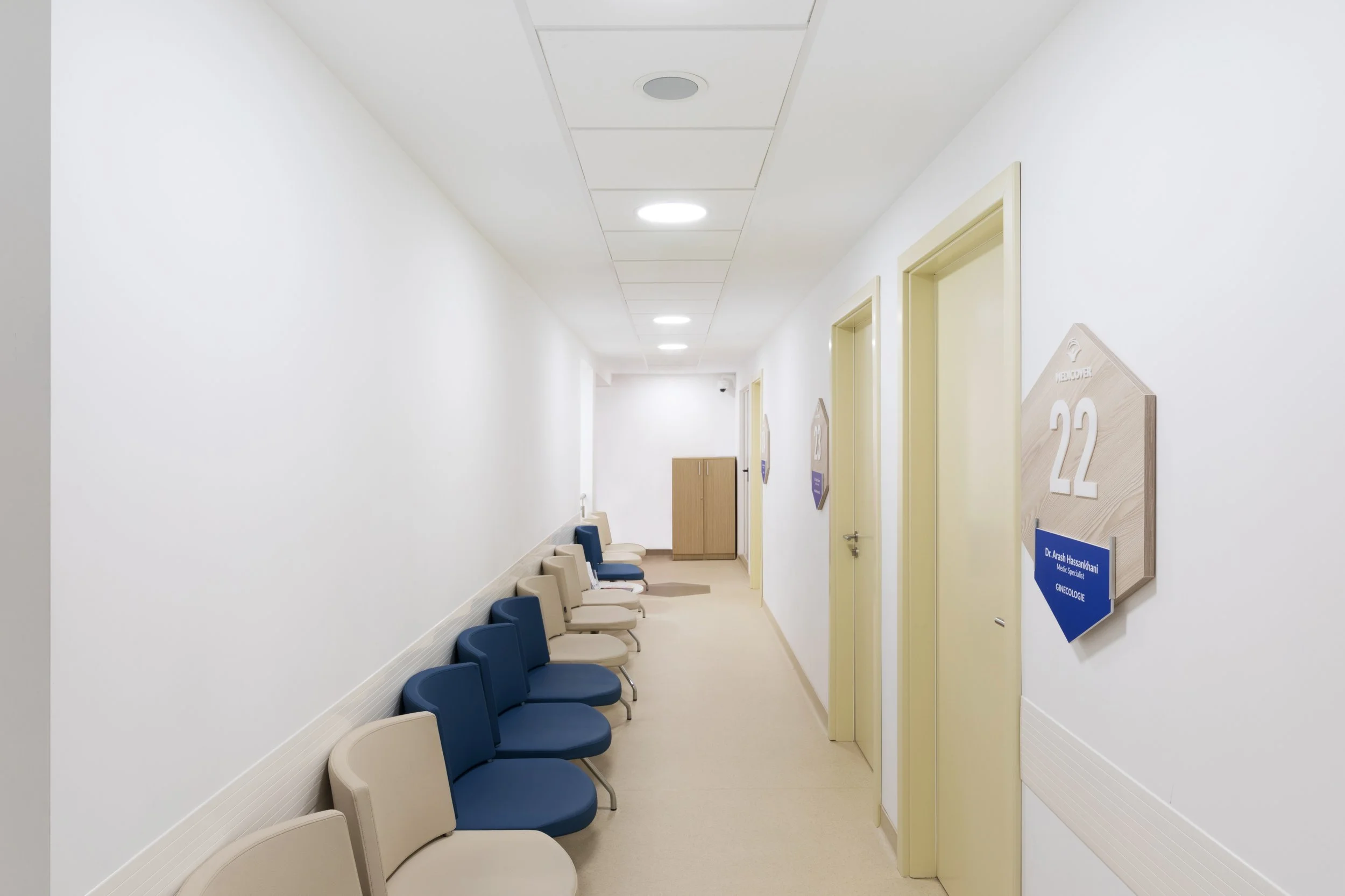 Empty hospital corridor with beige and blue chairs along the wall, closed doors, and signs indicating room numbers and medical departments.
