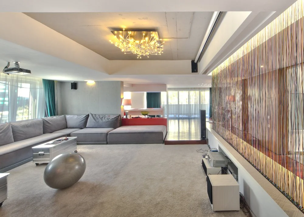 Modern living room with large gray sectional sofa, a metallic decorative ball on the carpet, and a wall-mounted flat-screen TV with a decorative wooden panel behind it. There is a large window with sheer curtains and teal drapes, a chandelier on the ceiling, and some furniture like a white cabinet and a red partition in the background.