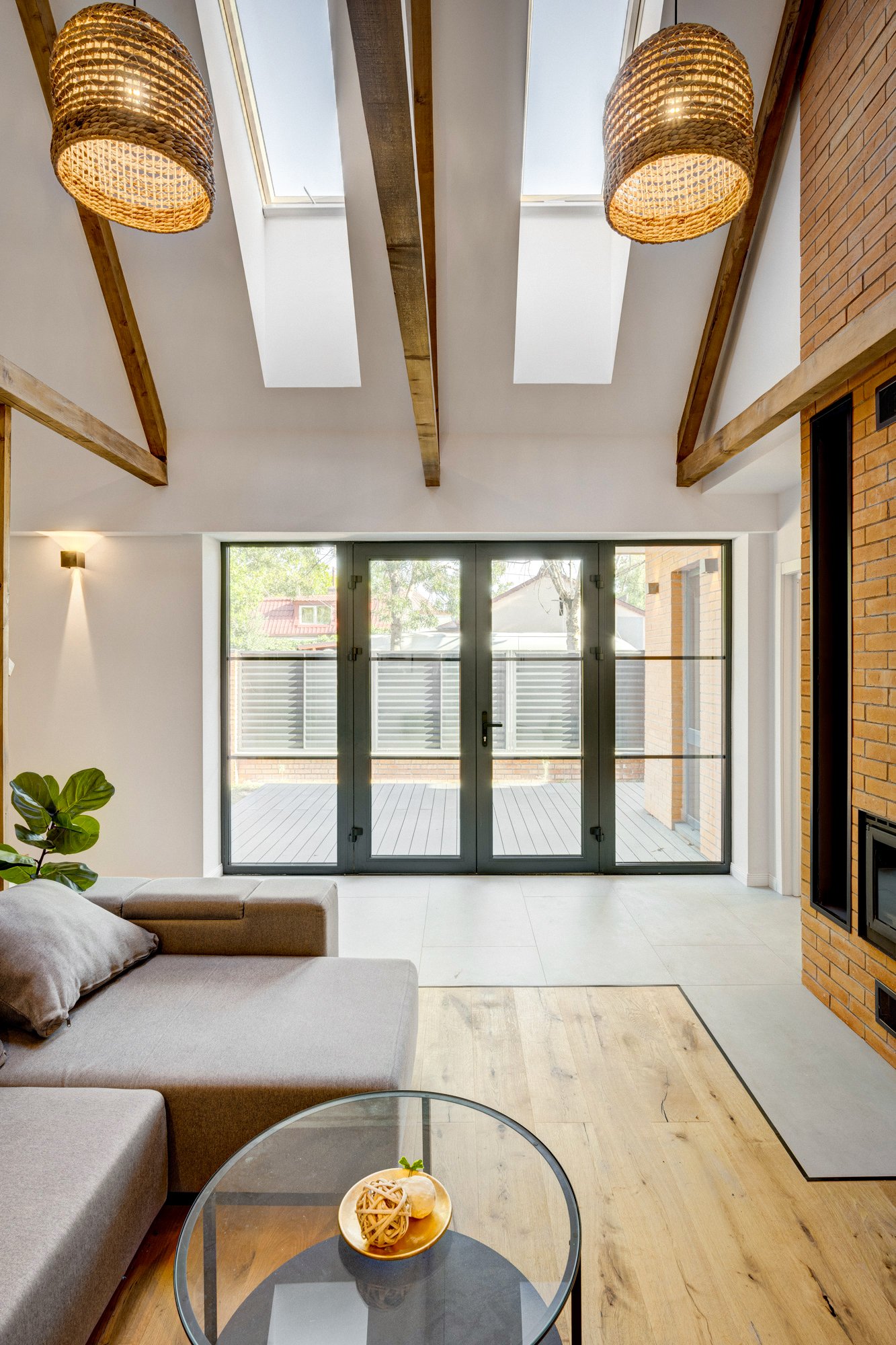 Living room with a beige sofa, a glass coffee table with a bowl of snacks, large glass doors leading to an outdoor patio, skylights, and a brick fireplace.