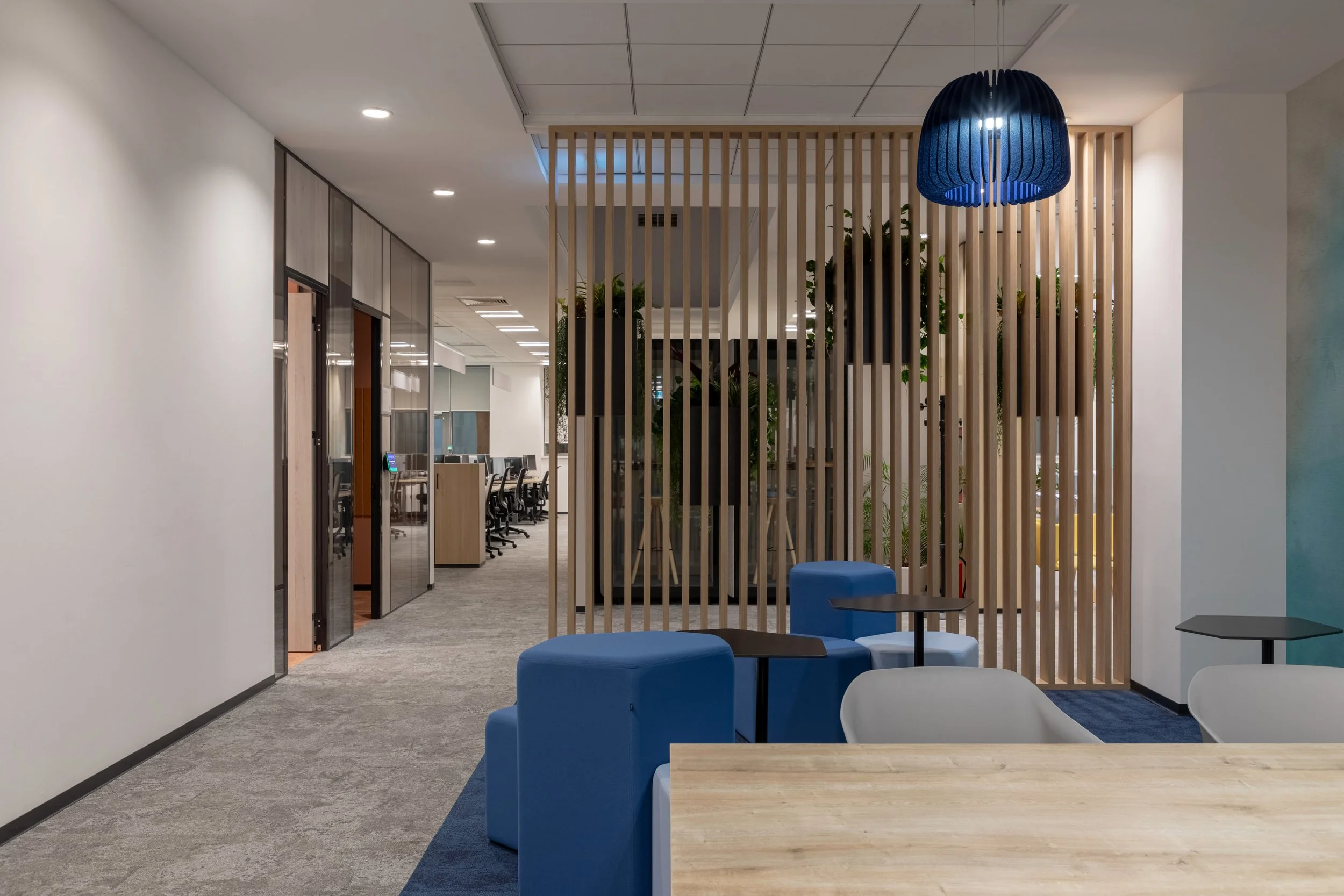 Modern office lounge area with blue and white chairs, small tables, wooden partition with plants, and an open workspace in the background.