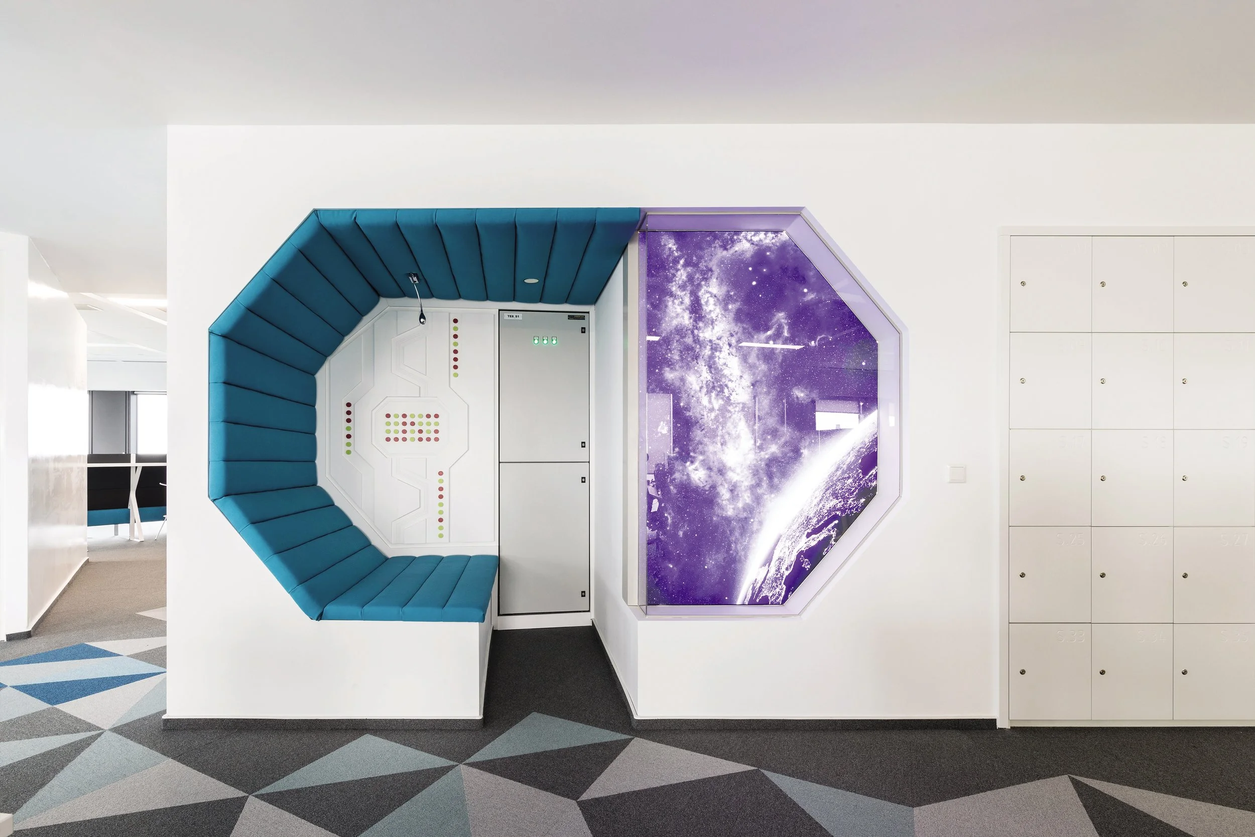 Modern office space with a blue, semi-circular seating area around a white wall with colored dot displays, and a large starry space image on a purple-tinted window. White lockers are on the right and a patterned floor is below.