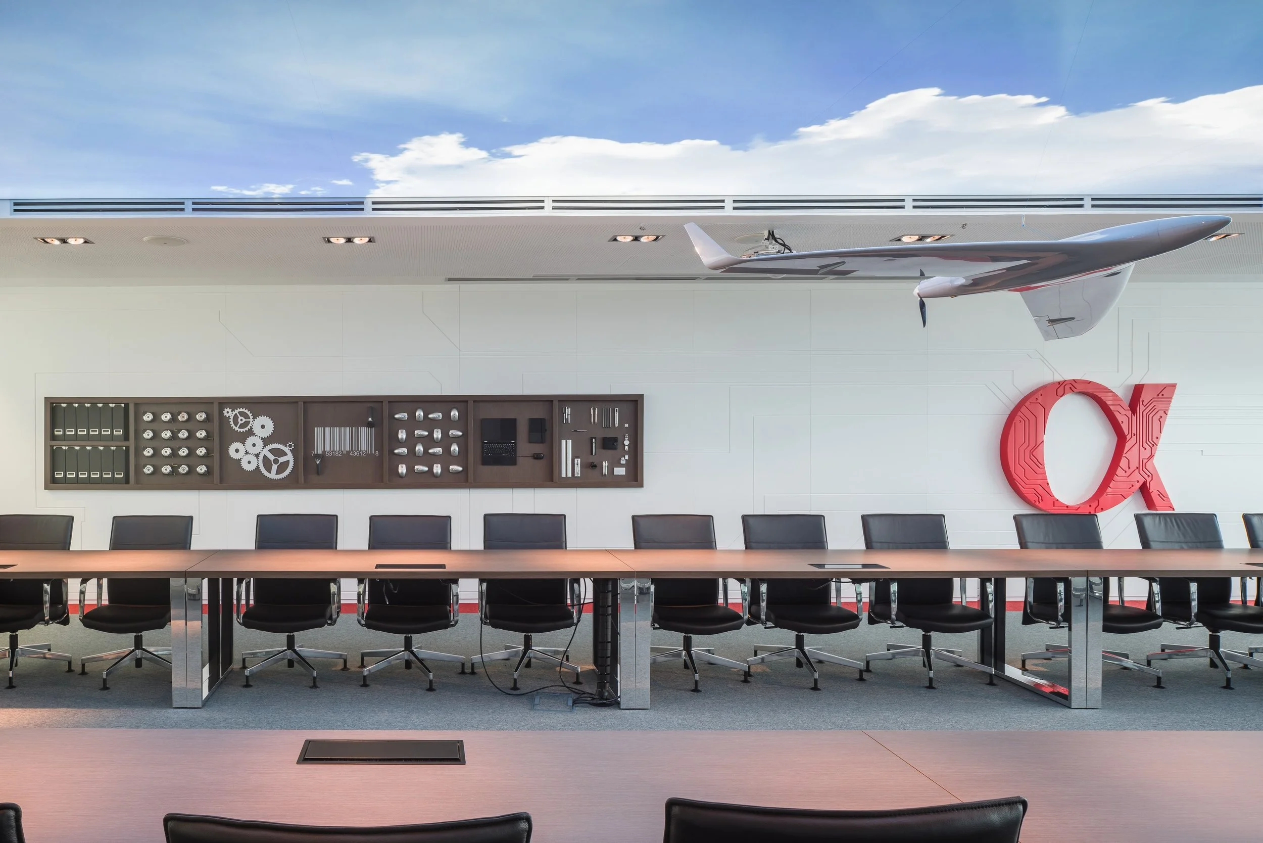 A conference room with a long wooden table, black chairs, a wall-mounted display of mechanical parts, a large red 'QX' logo, and an airplane model hanging from the ceiling against a sky mural.