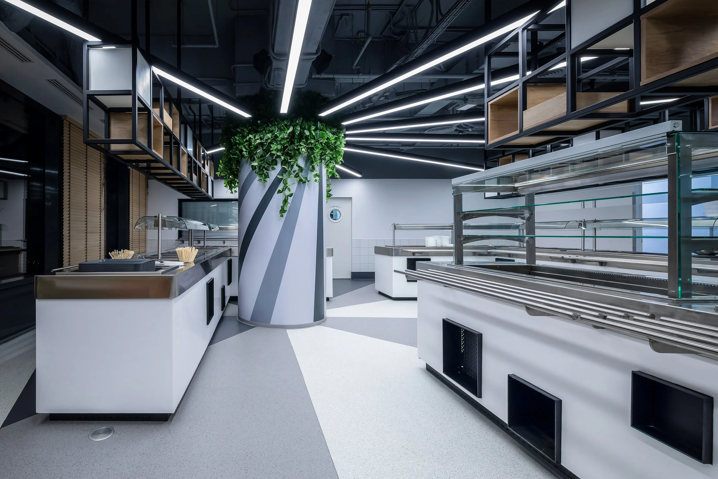 Modern cafeteria or food service area with stainless steel counters, including a food display and prep station, modern black and white ceiling design with linear lighting, a large decorative column with green plants, and clean, minimalist design.