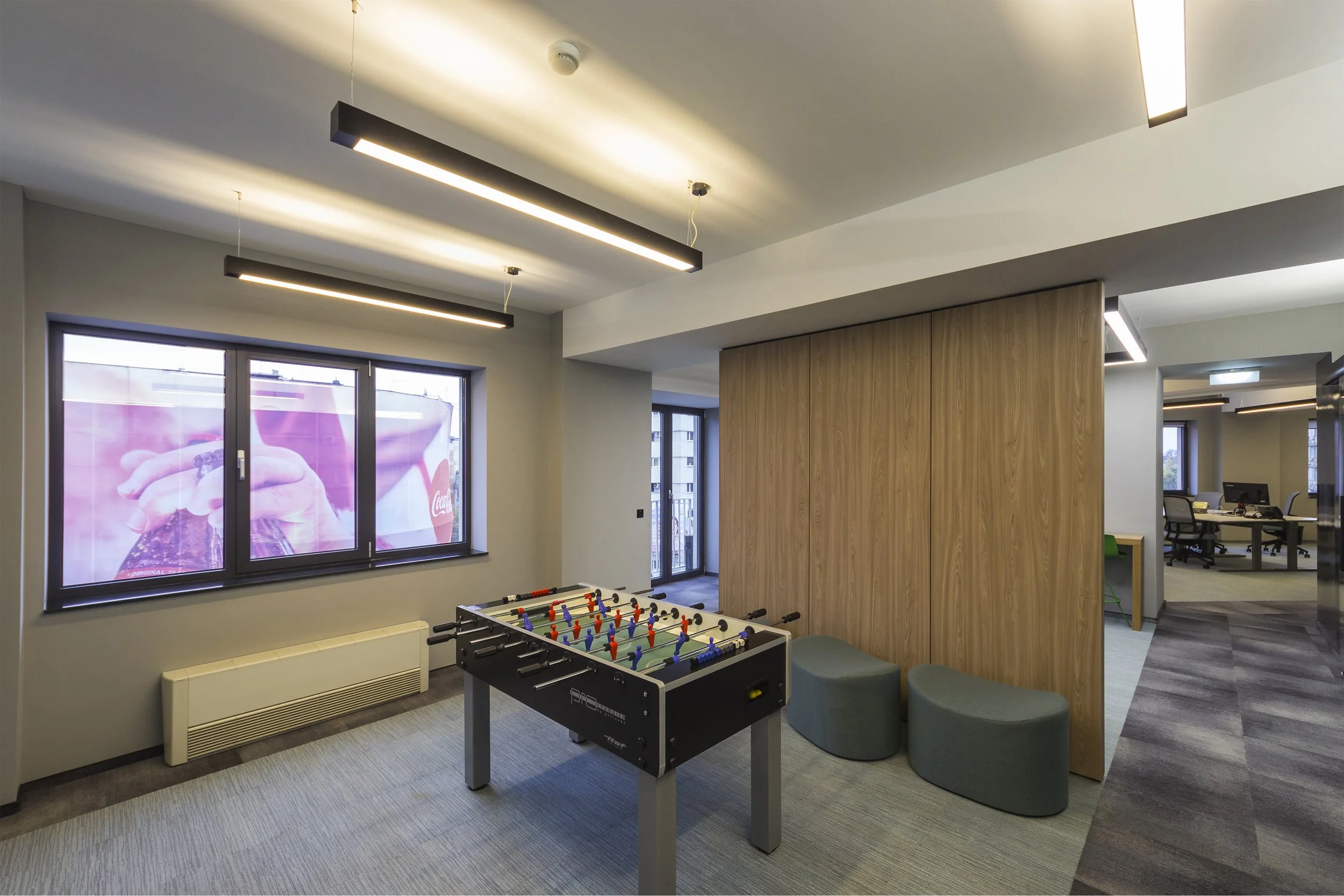 Modern office lounge area with a foosball table, two gray seating poufs, large windows, and ceiling-mounted lighting fixtures.