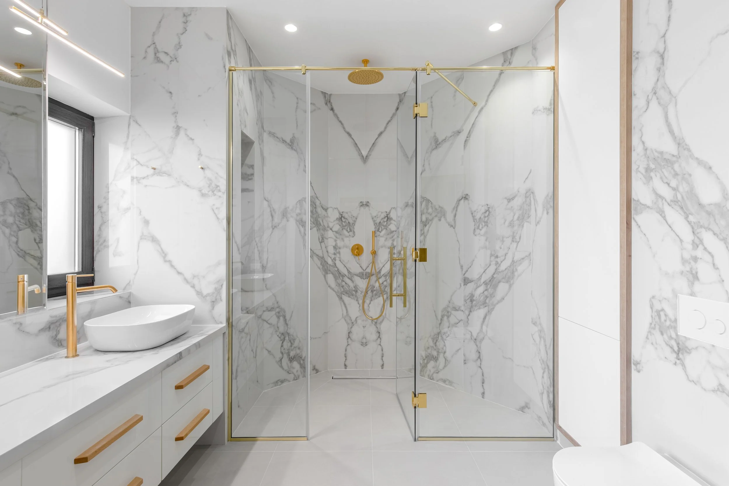 Modern bathroom with white marble walls, a glass shower with gold fixtures, a white vanity with a vessel sink, and a mirror.