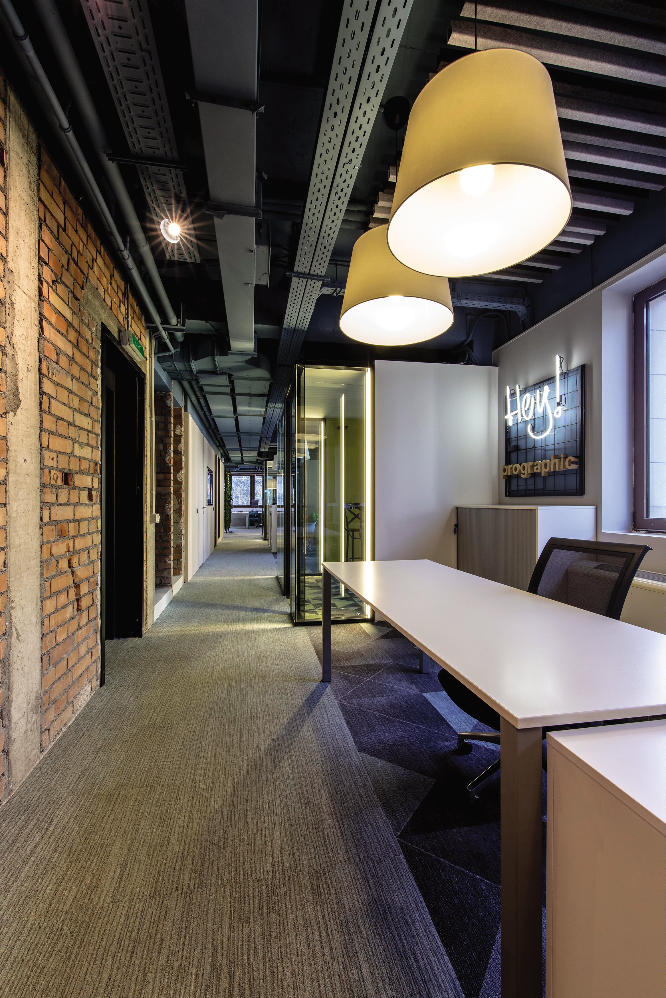 Modern office hallway with exposed brick wall, ceiling with visible piping, hanging oval lamps, glass enclosure, and wall sign that says 'Hey!'.