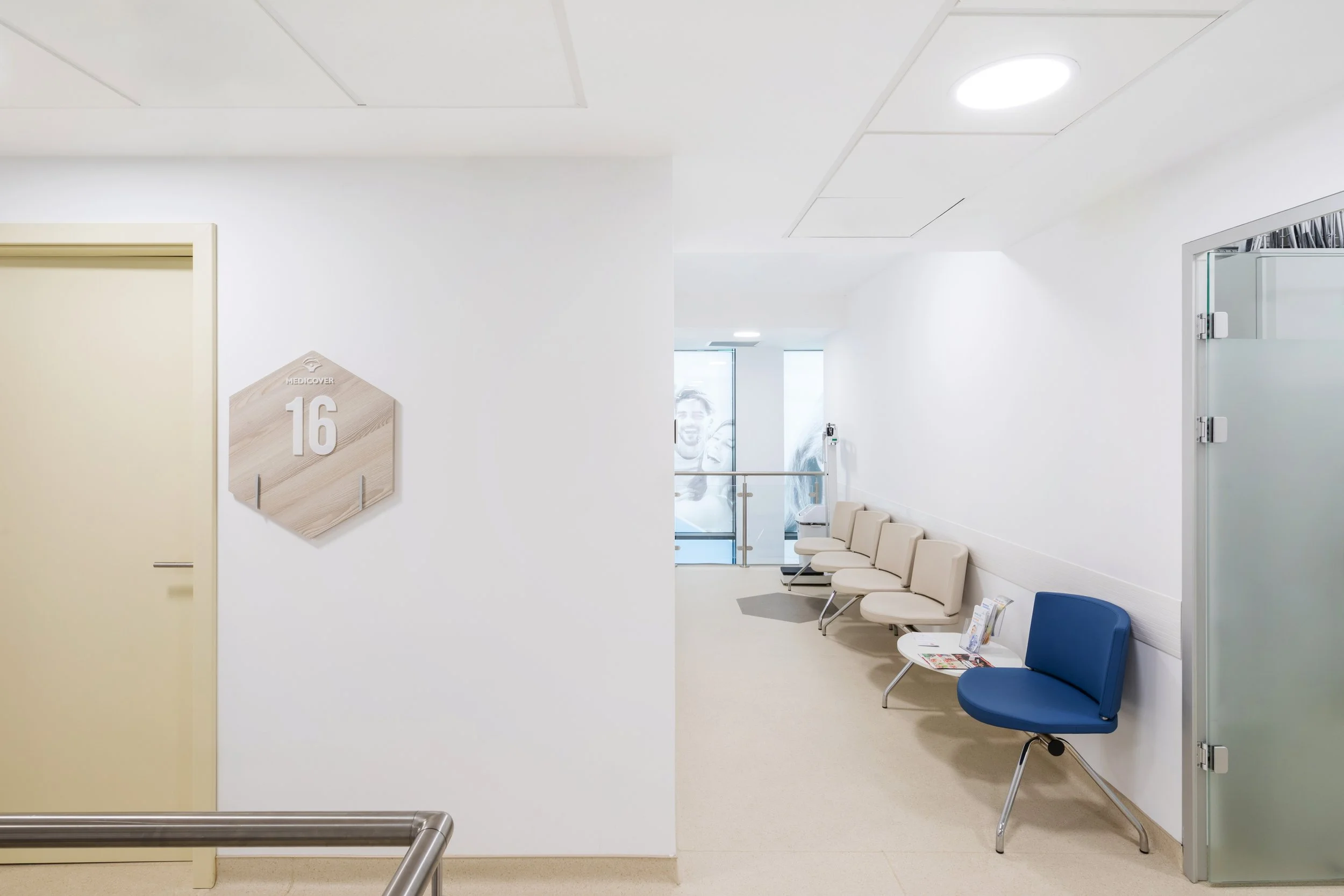 An empty hospital waiting room with beige chairs, a blue chair, magazines on a small white table, a white wall with a wooden hexagon sign with the number 16, and frosted glass door.