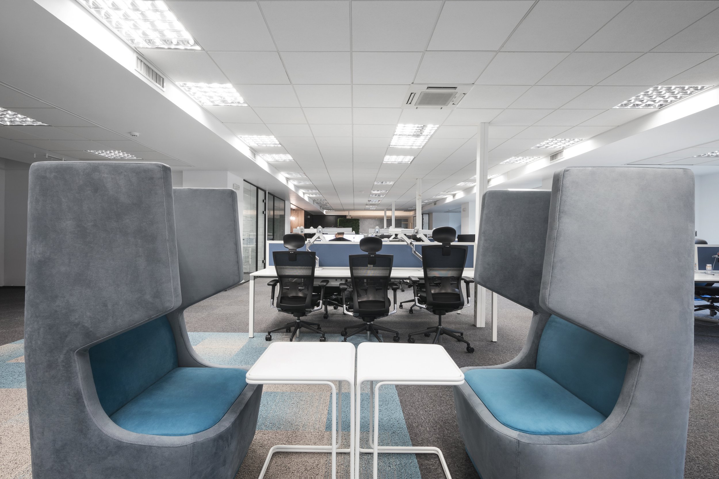 Interior view of a modern office with unique high-back seating booths in the foreground, separated by small white tables, and multiple workstations with chairs in the background.