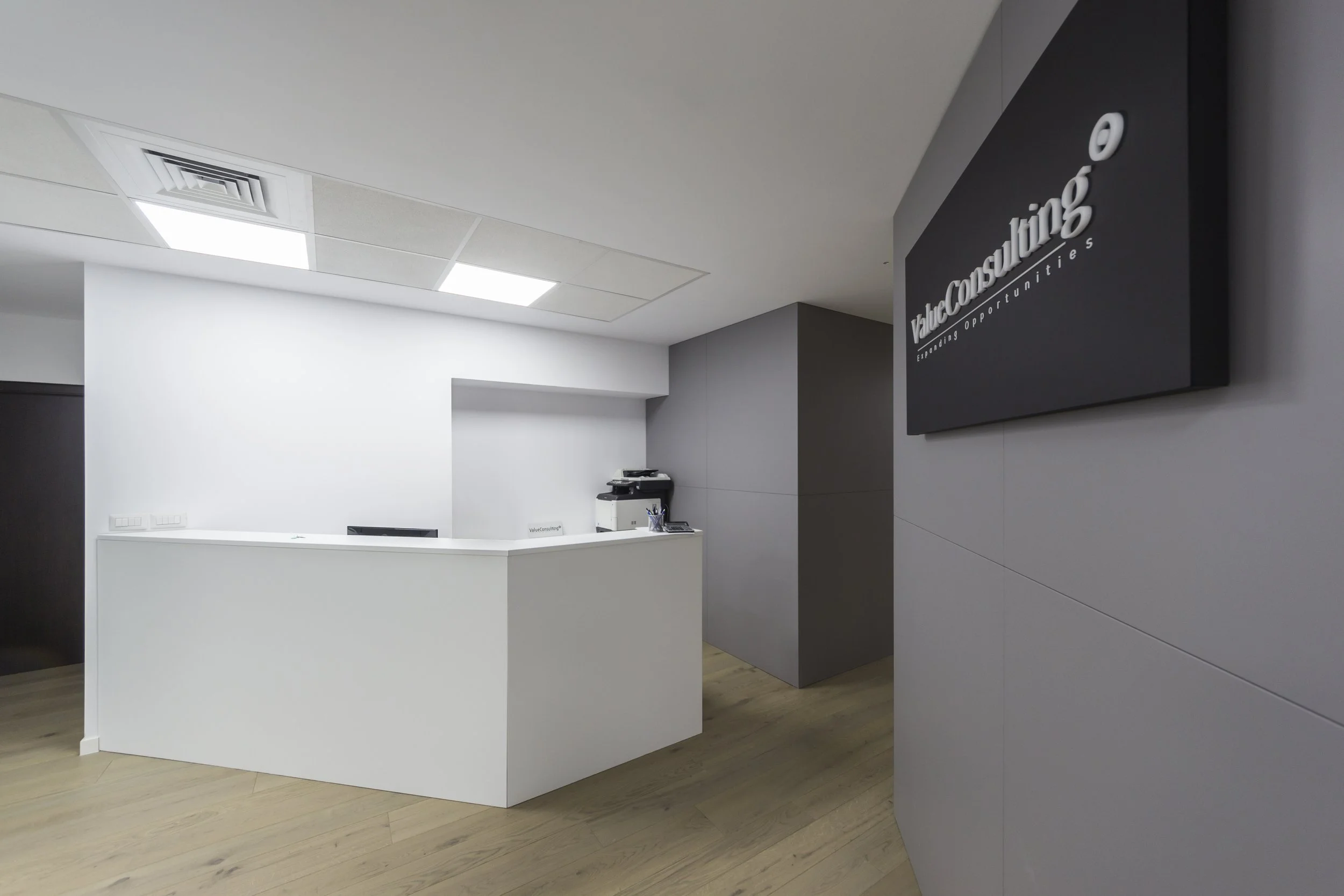 Office reception area with a white curved desk, walls painted in white and gray, and a mounted black sign reading 'Value Consulting' in white letters with slogan 'Empowering Opportunities'.