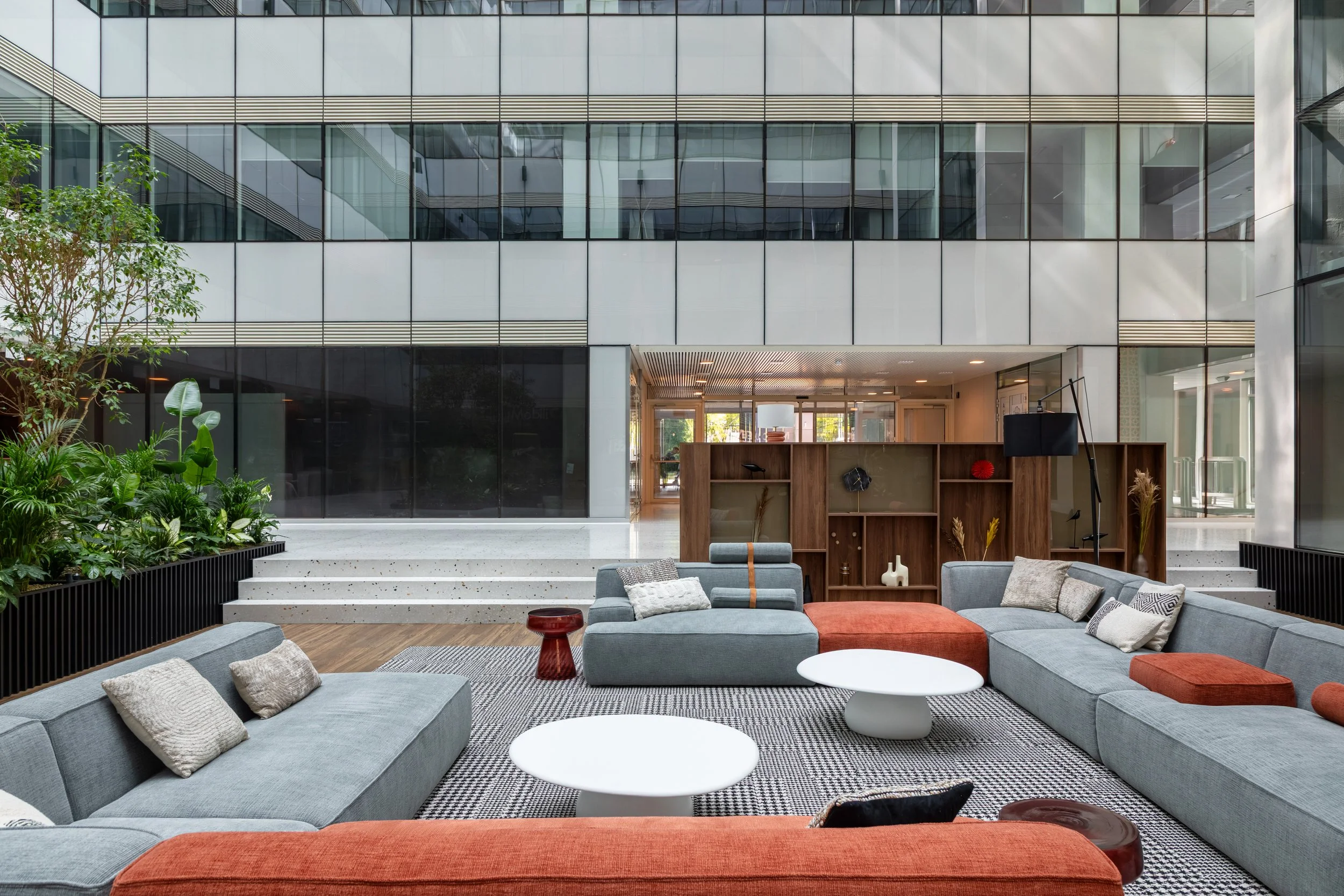 Modern office lobby with gray and orange upholstered sofas, white coffee tables, a wooden bookshelf with decorative items, indoor plants on the left, and large glass windows and doors in the background.