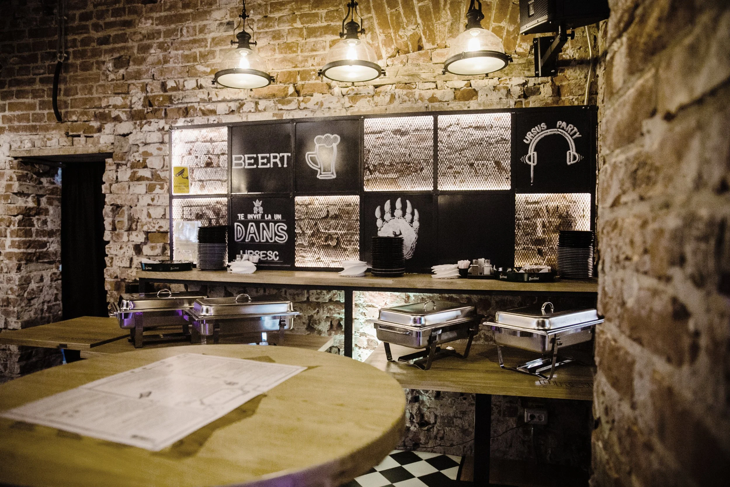 Bar area with exposed brick walls, black chalkboard signs with beer and party drawings, stacked black plates, napkins and condiments, and chafing dishes on a wooden table in a rustic restaurant.