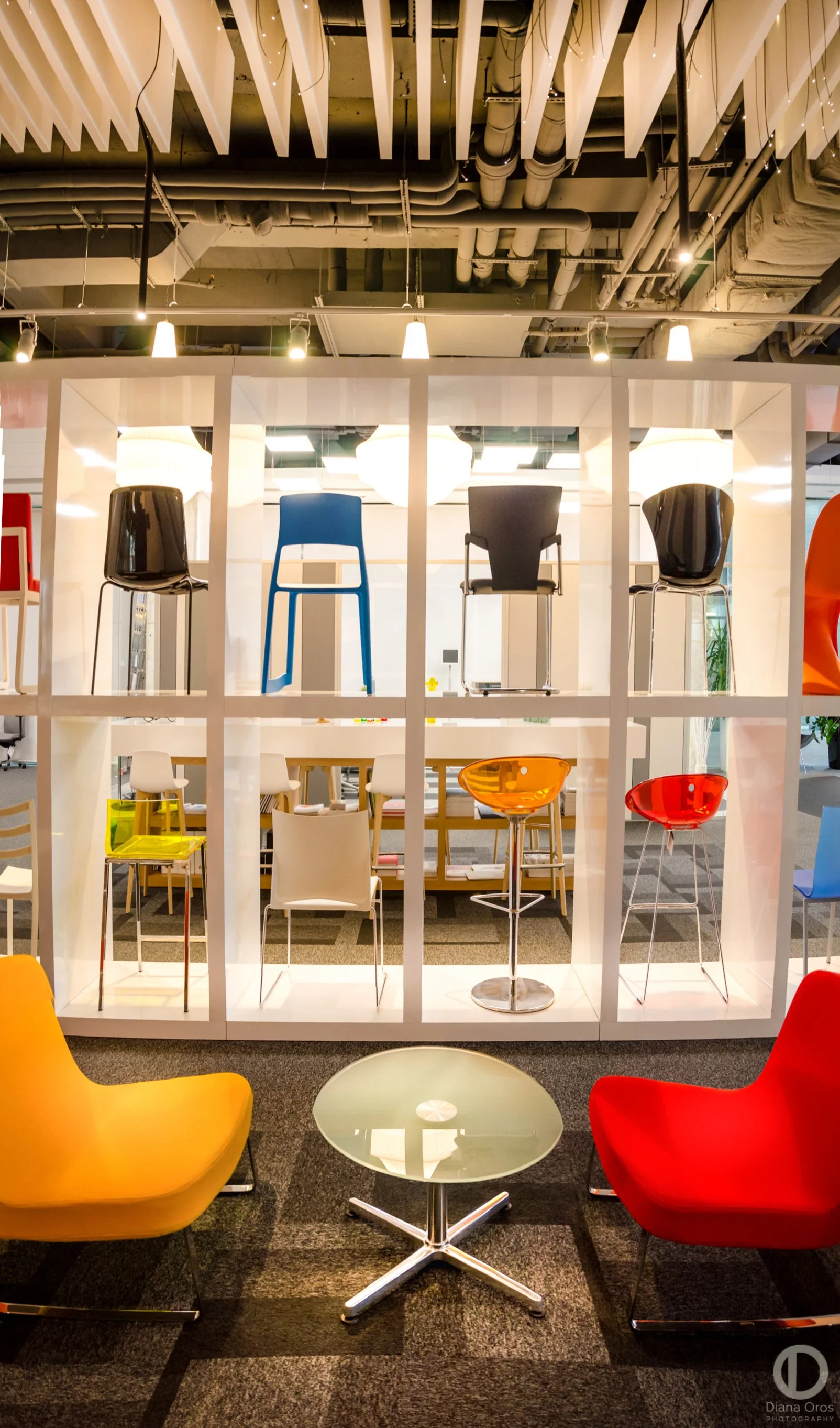 Display of colorful chairs and barstools in a modern office or showroom with open ceiling and industrial lighting.
