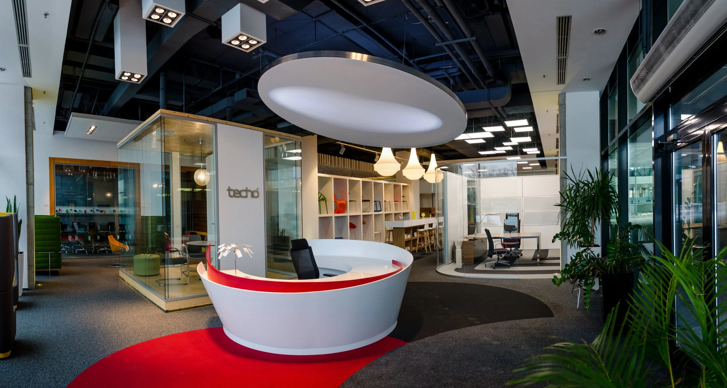 Modern office reception area with white curved desk, large oval ceiling light, glass enclosed meeting room with chairs, bookshelf with colorful decor, and potted plants near the entrance.