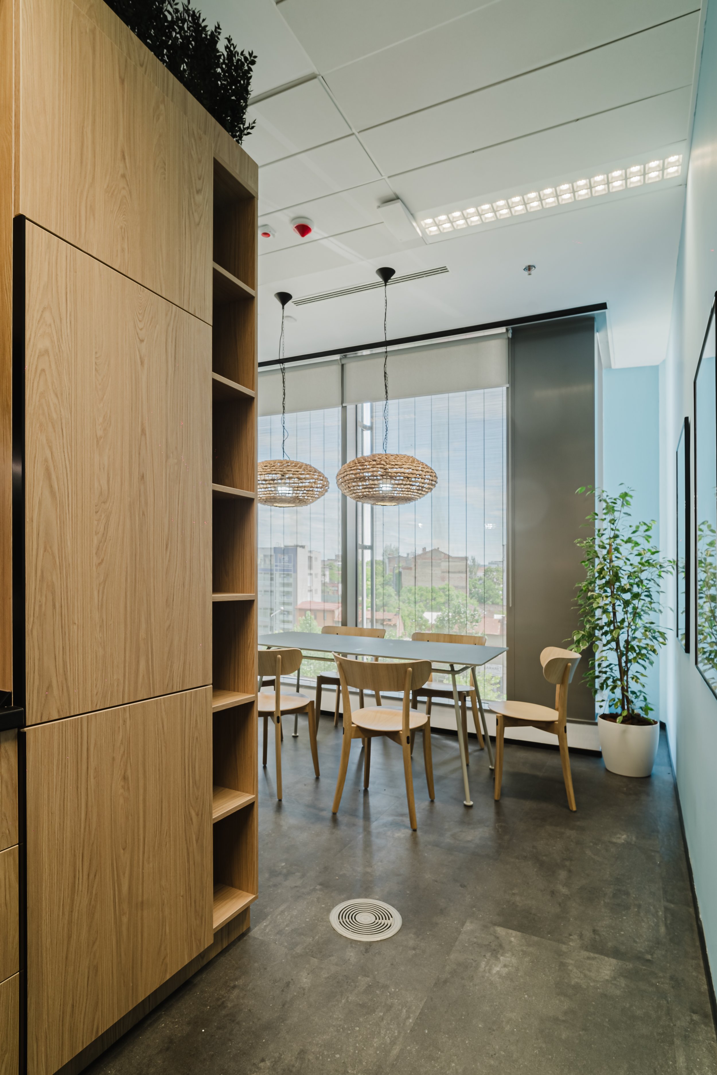 A modern conference room with a wooden cabinet on the left, a table with six wooden chairs, large windows with blinds, hanging wicker light fixtures, a potted plant, and framed artwork on the wall.