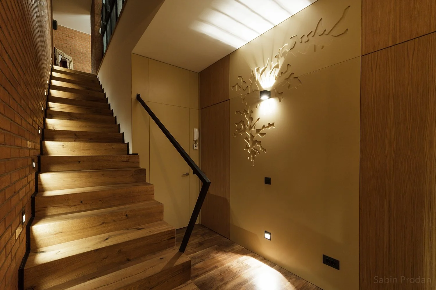Interior view of a staircase with wooden steps, a black handrail, and a decorative wall light casting a pattern on the beige wall. The walls are a mix of brick and wood paneling, with small floor lights illuminating the space.