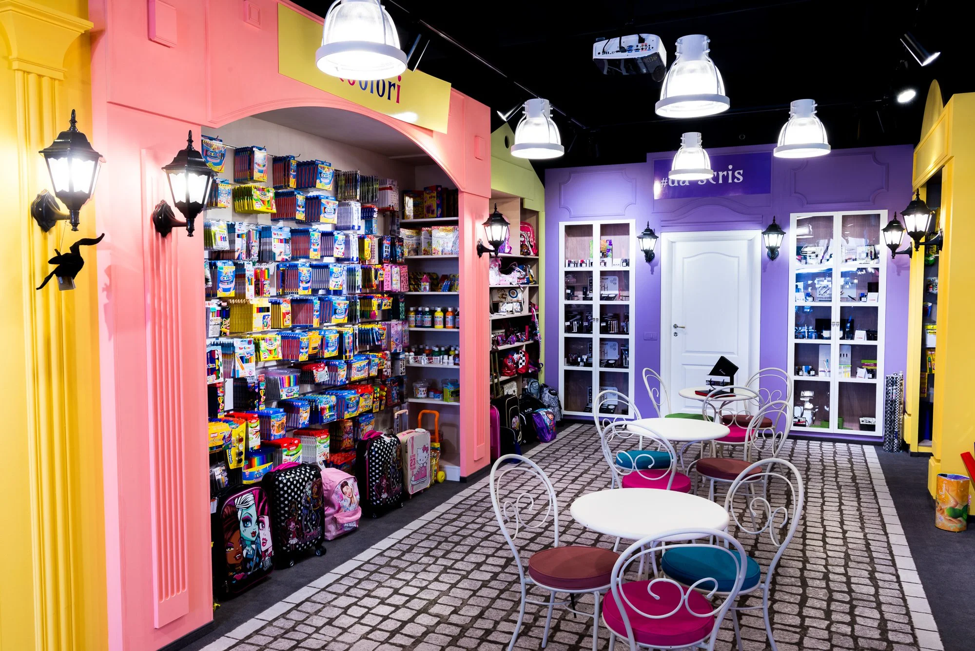 Colorful room with shelves filled with school supplies, backpacks, and toys, white tables with ornate chairs, and purple and yellow walls with decorative lighting.