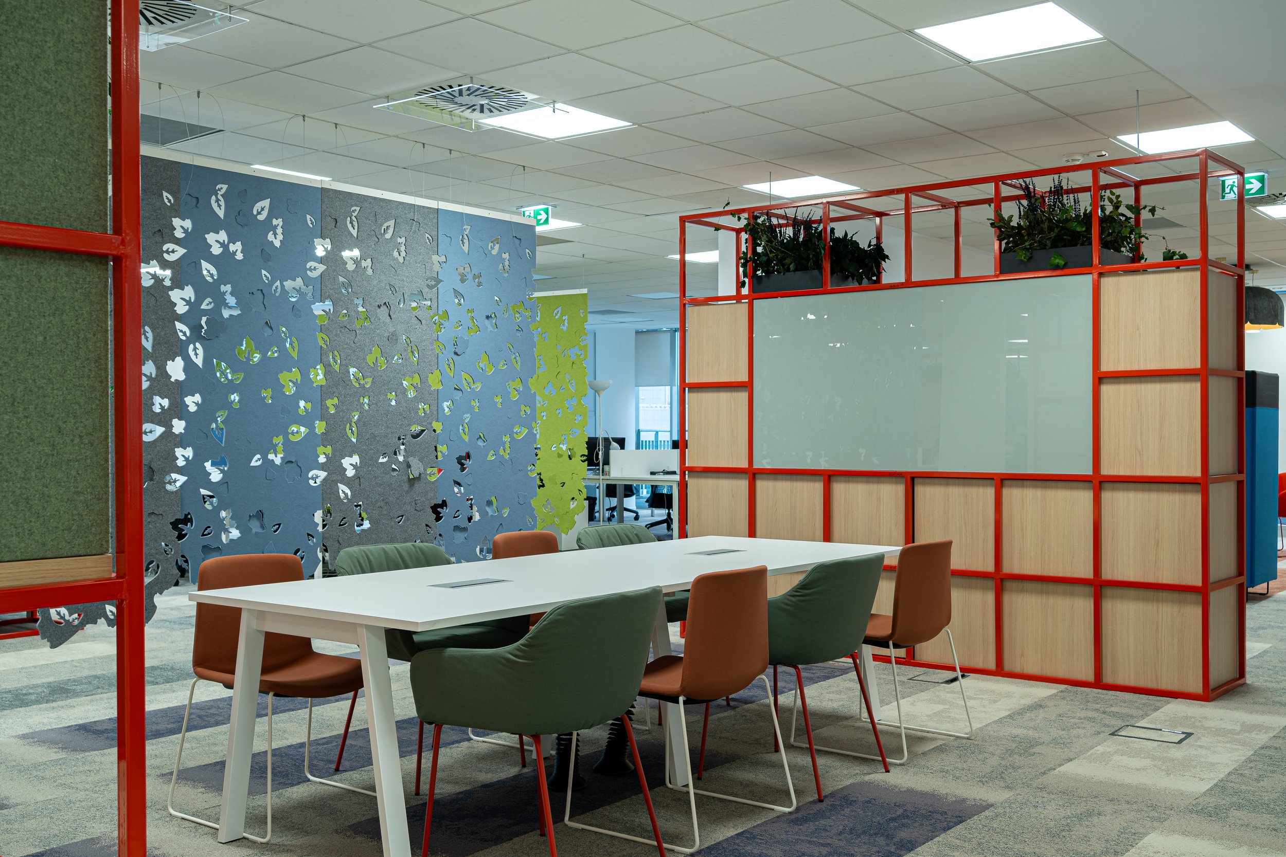 Modern office conference room with a white table and six chairs, colorful partition panels, and decorative plants on a raised platform, with ceiling lights and exit signs.