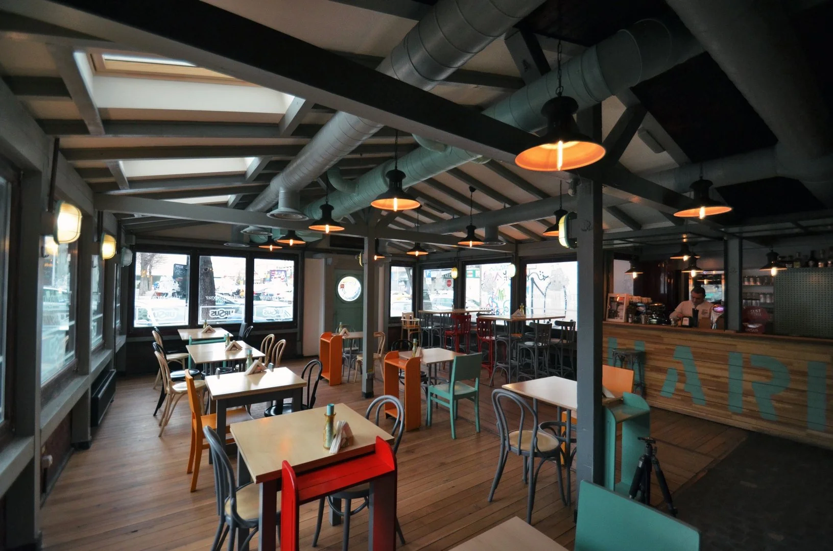 Interior of a modern cafe with wooden floors, colorful chairs, and tables. Large windows let in natural light, with a barista working behind the counter.