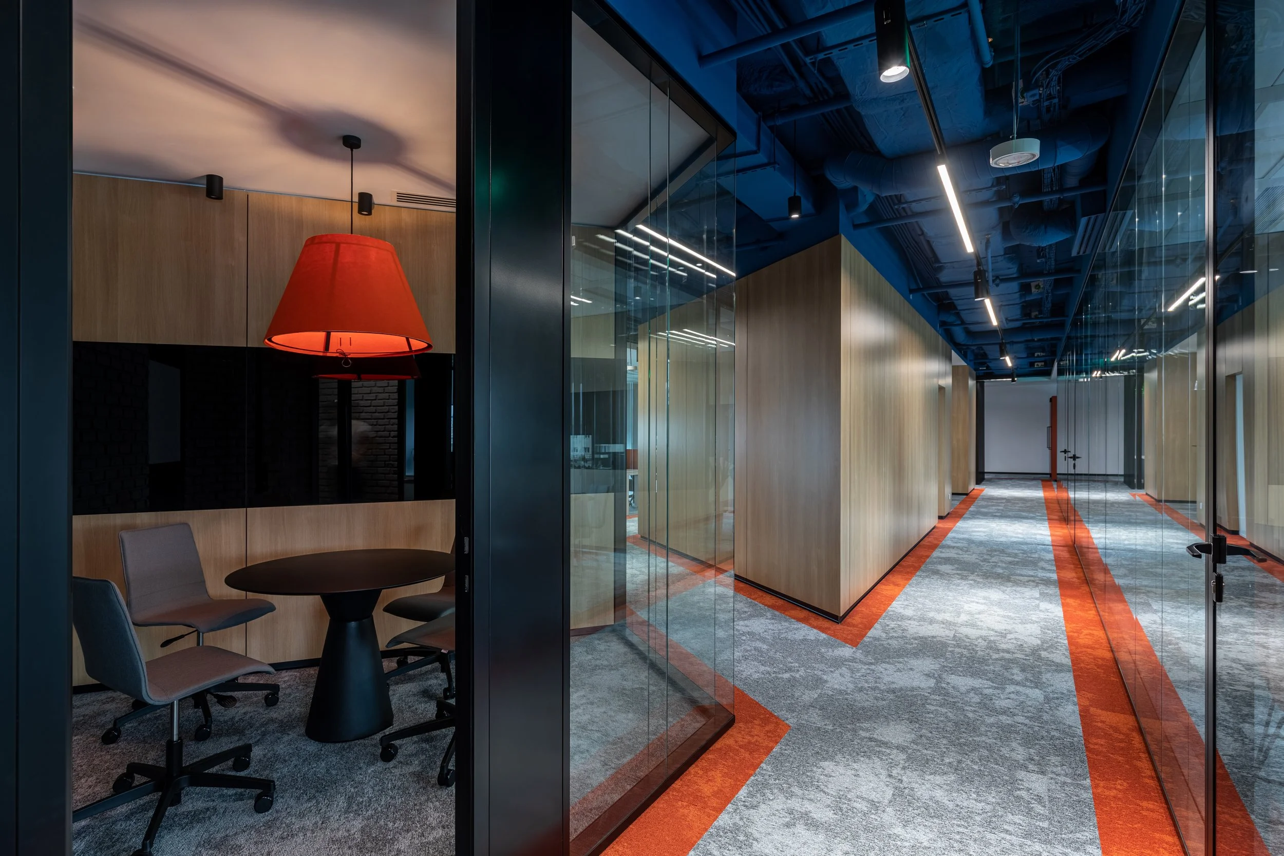 Modern office corridor with glass walls, wood paneling, grey and orange carpet, and blue ceiling. There is a small conference room with a round table, chairs, and a red hanging lamp.