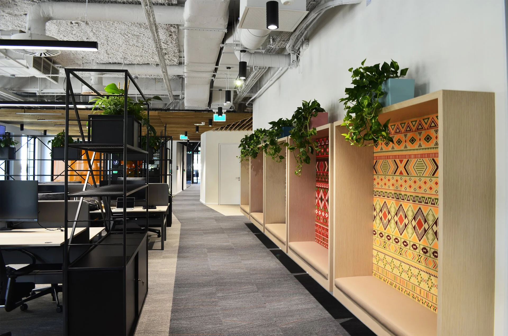 Modern office space with workstations, plants, and decorative partition booths with patterned fabric backs along a hallway.