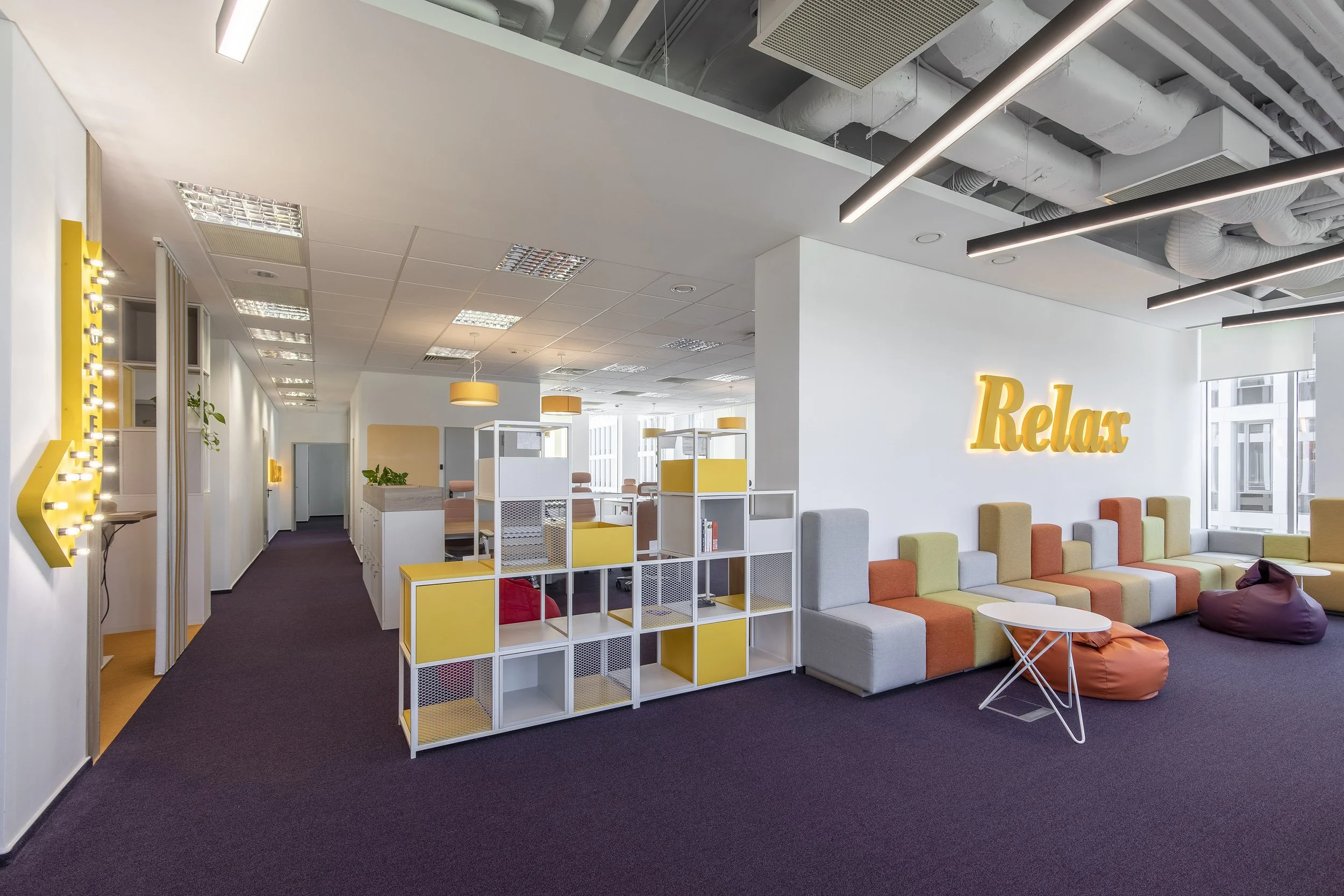 Modern office lounge with colorful seating, a white wall with orange illuminated "Relax" sign, and decorative shelving with yellow accents.