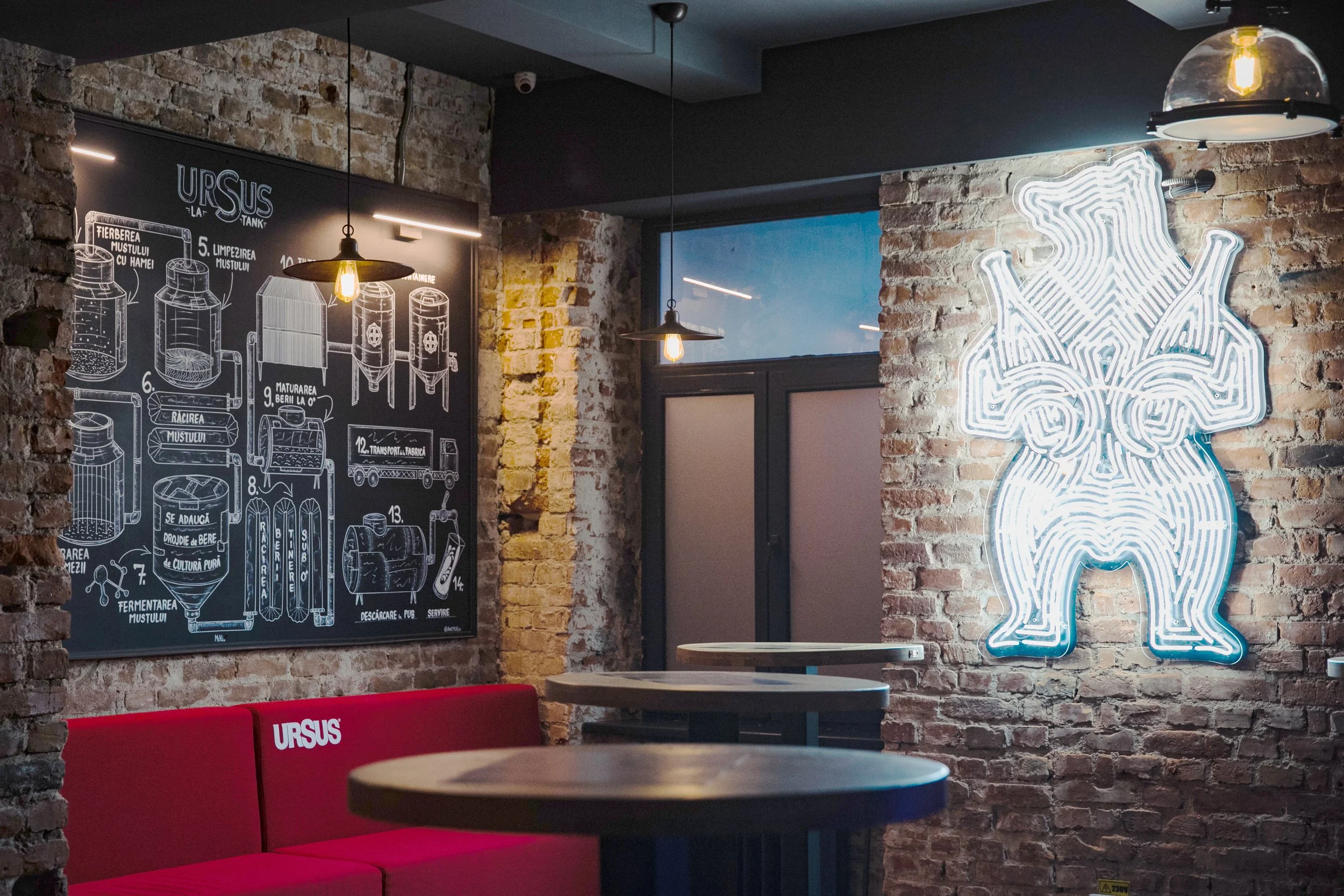 Interior of a modern brewpub with exposed brick walls, a neon sign of a rabbit, a chalkboard with brewing instructions, and a seating area with red benches and round tables.