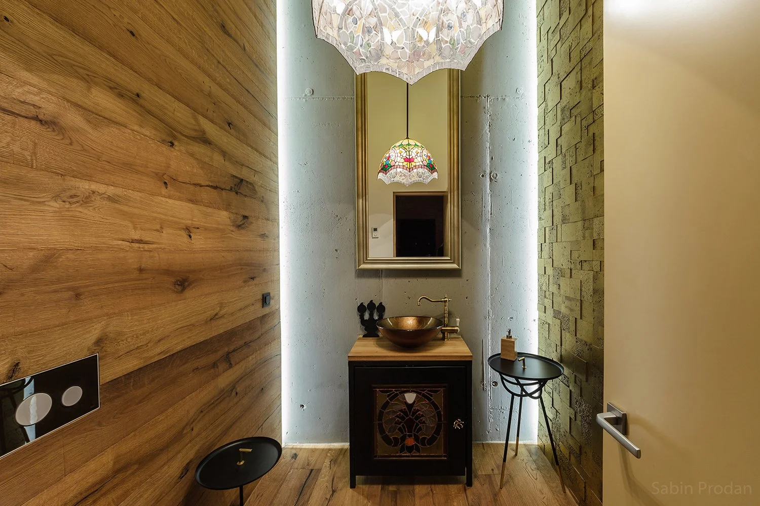A modern bathroom with wooden wall on the left, green textured wall on the right, and concrete wall behind. Contains a small black cabinet with ornate front, a copper basin sink, a gold faucet, and a large mirror framed in gold. Two hanging stained glass lamps are reflected in the mirror, and there are two small black stools in front of the sink.