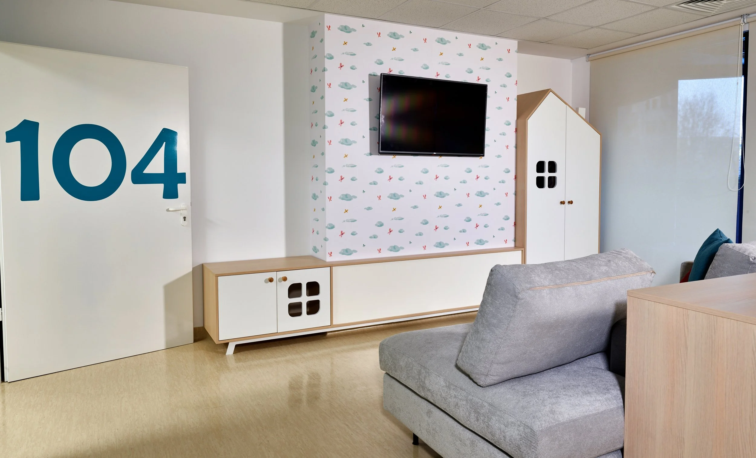 Hotel room with the door numbered 104, a wall-mounted TV, a gray sofa, a white and wood storage cabinet, and a large window with a roller blind.