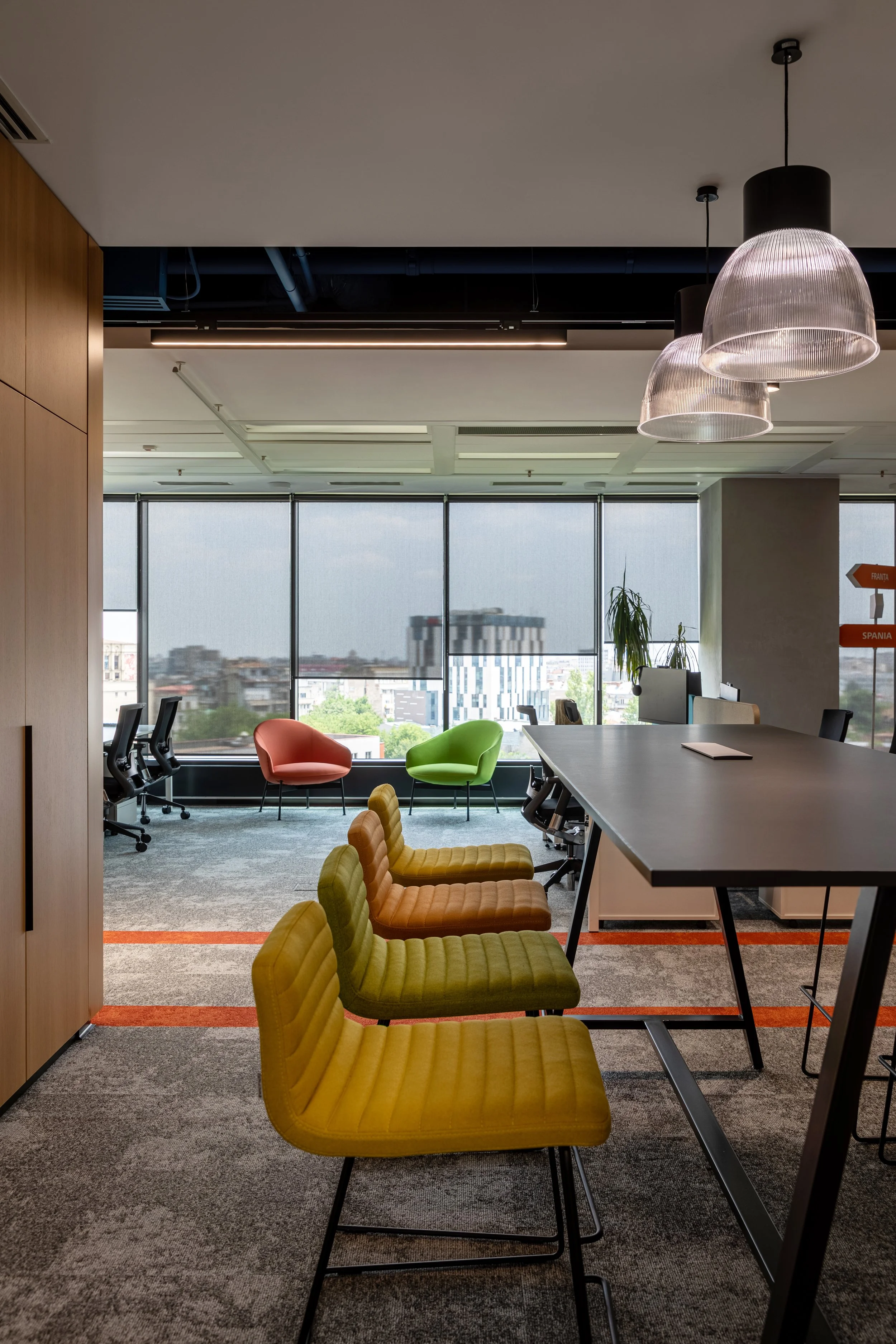 Modern office space with colorful chairs, large windows with city view, and hanging pendant lights.