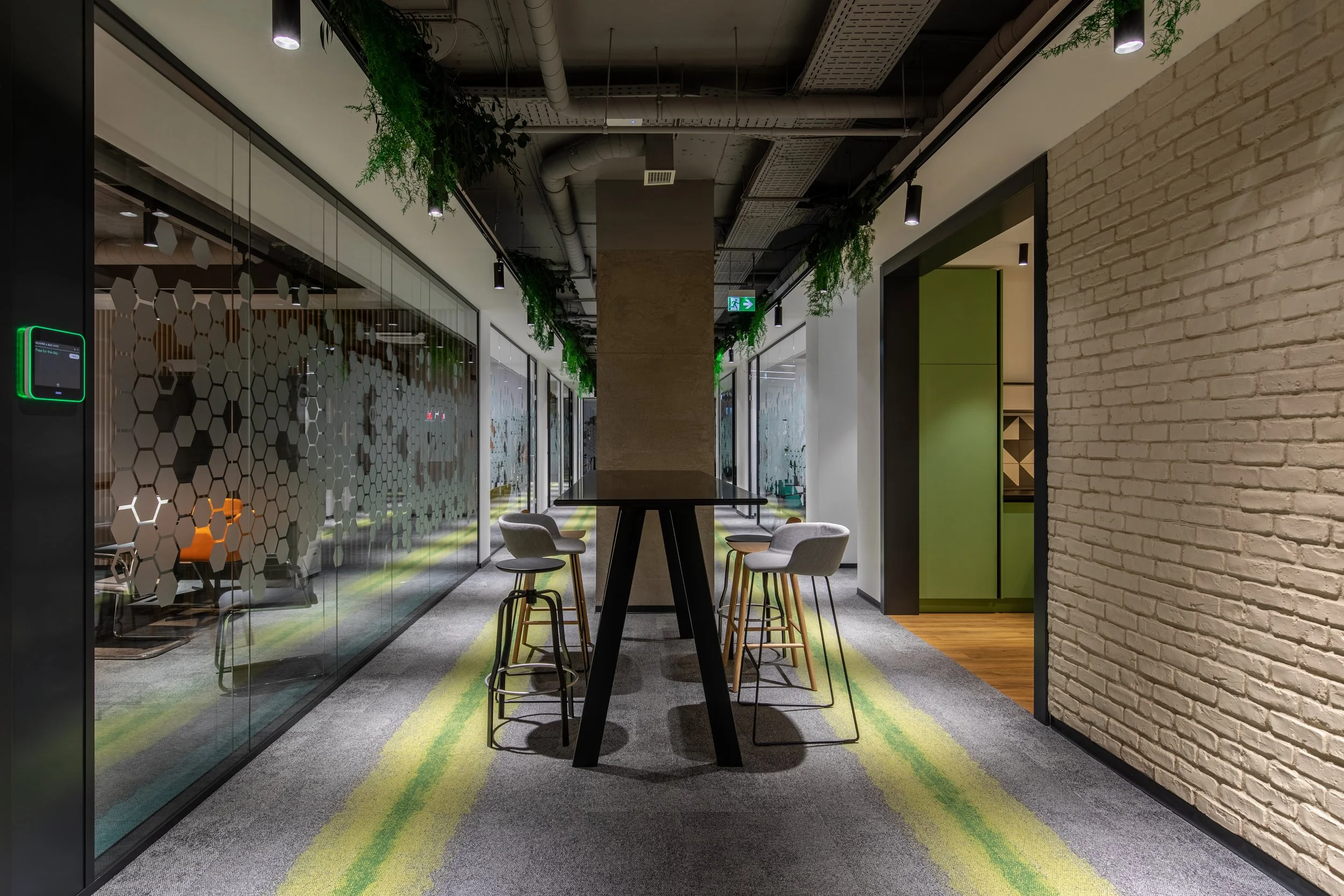 Modern office corridor with glass partitions on the left, a high table with seating in the center, green plants hanging from the ceiling, brick wall on the right, and an exit sign overhead.