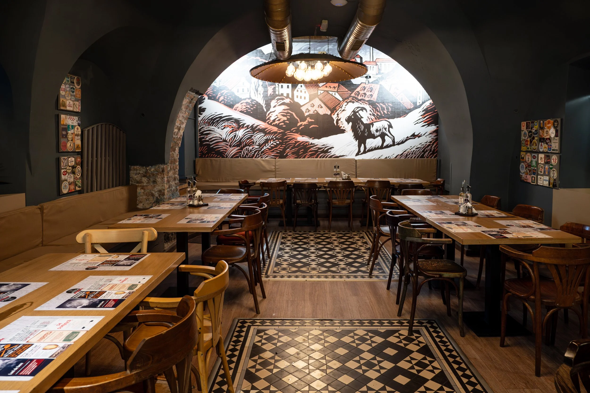 Empty restaurant with wooden tables and chairs, artistic mural of a mountain landscape with a lion on the back wall, and patterned black and white floor tiles.