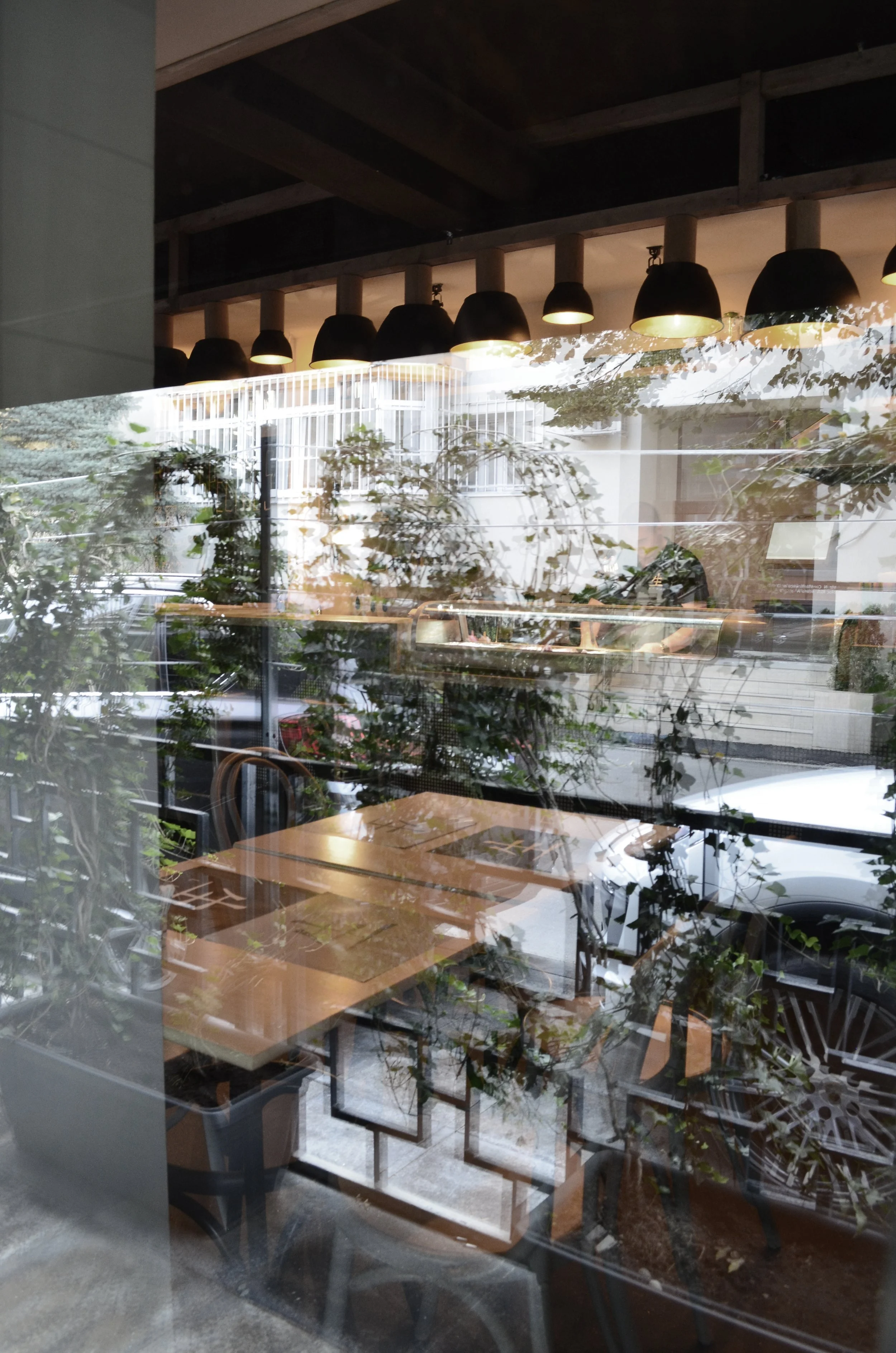 Inside view of a cafe or restaurant with wooden tables, chairs, and modern black hanging lamps, seen through a glass window that reflects outdoor trees and street scene.