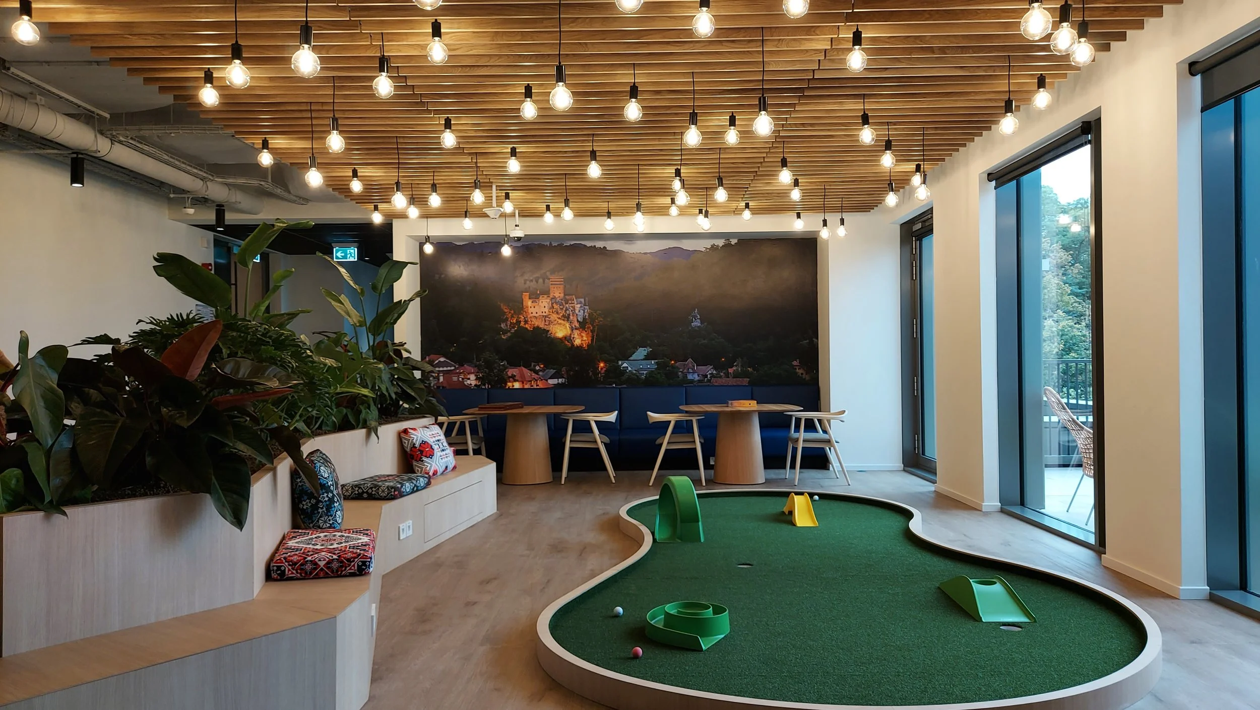 Indoor office lounge area with a mini golf putting green, wooden ceiling with hanging light bulbs, large window with outdoor balcony, green plants, and a mural of a castle on the back wall.