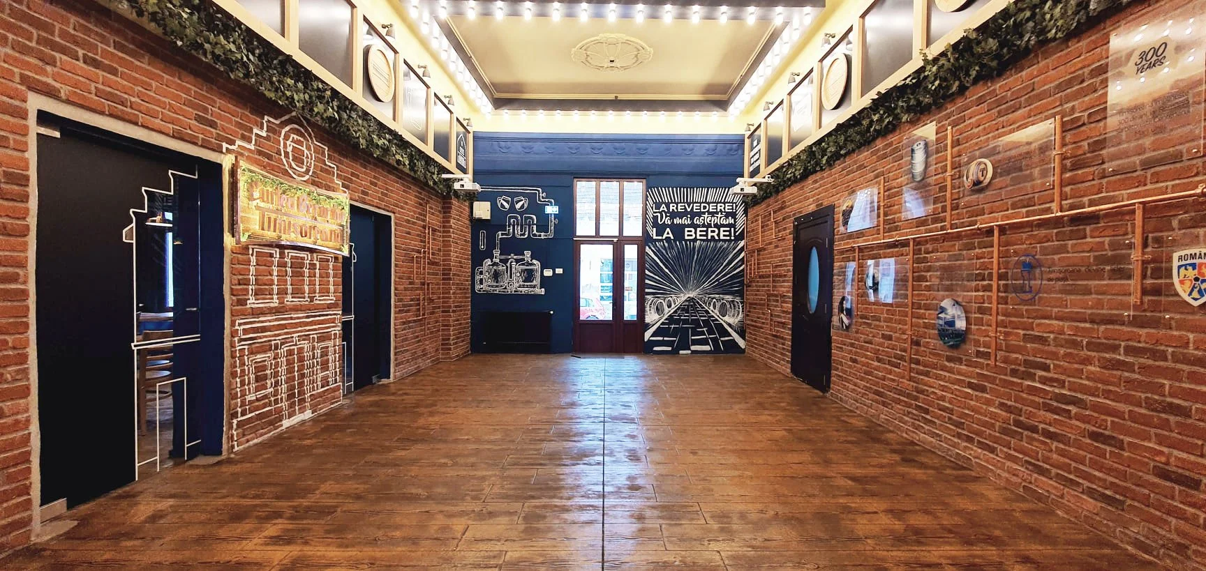 Interior of a room with brick walls, wooden floors, and blue accents. There are framed photos and artwork on the walls, including a mural with the words "LA REVEDEER! VAMAI GESTIUN LA BERE!". The ceiling has decorative molding and lighting. The room appears to be part of a bar or brewery.