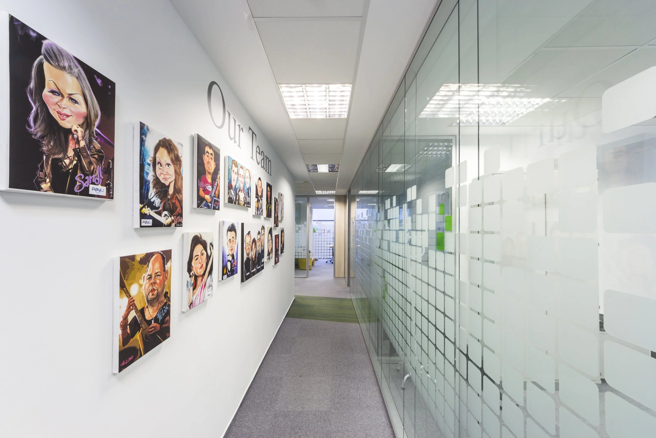 Office hallway with caricature portraits of team members on the wall and a glass-walled meeting room on the right, with the words 'Our Team' on the wall.