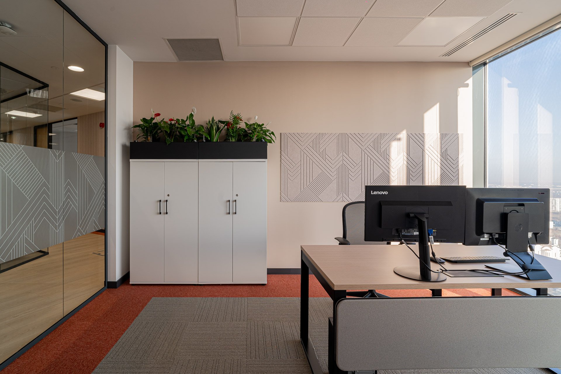 An office with a desk, dual computer monitors, a chair, a white cabinet with plants on top, a wall with patterned decor, and a large window showing a cityscape.