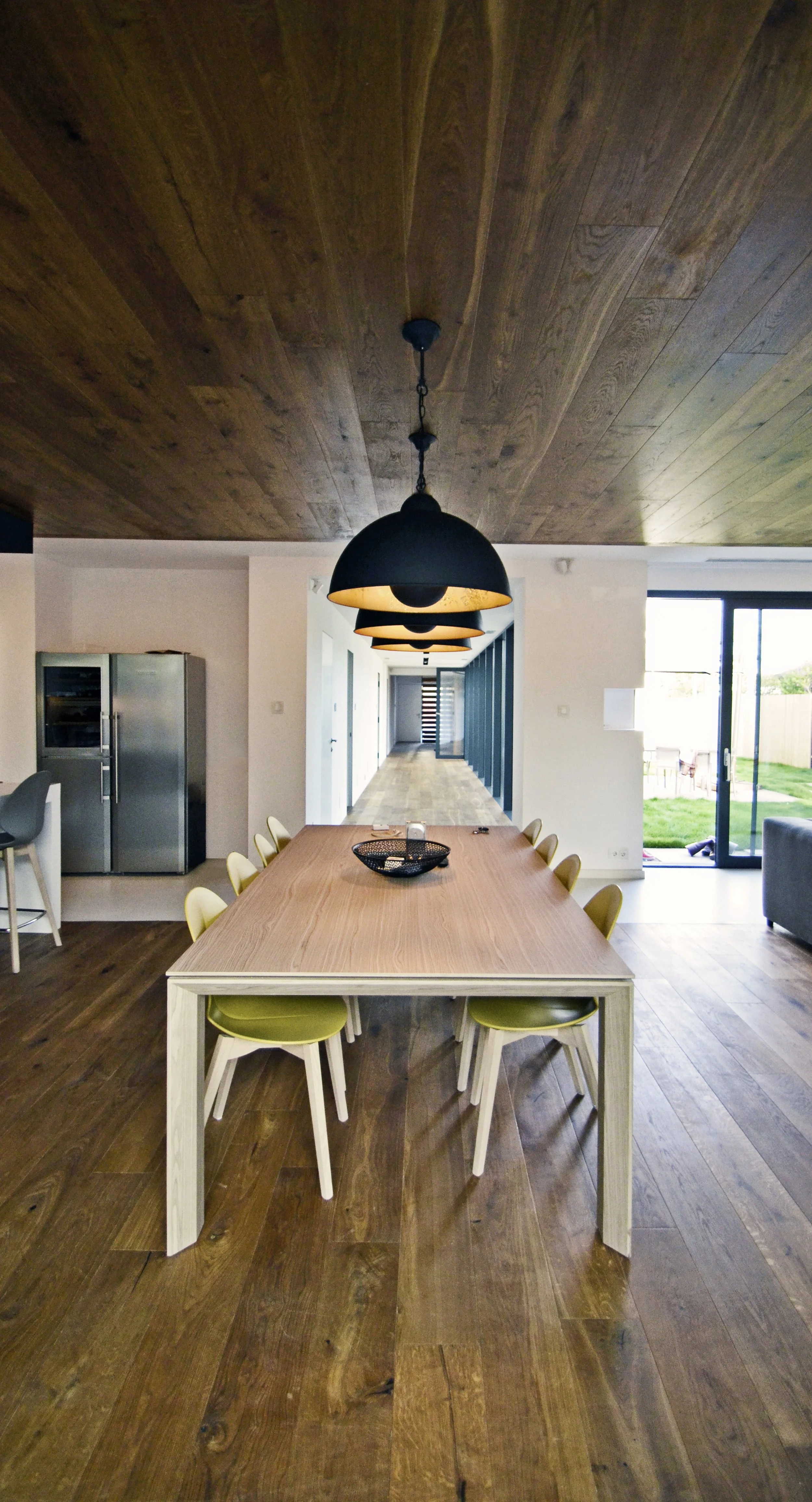 A modern dining room with a long wooden table, yellow chairs, black hanging pendant lights, and a view of a hallway and sliding glass door leading to a grassy yard.