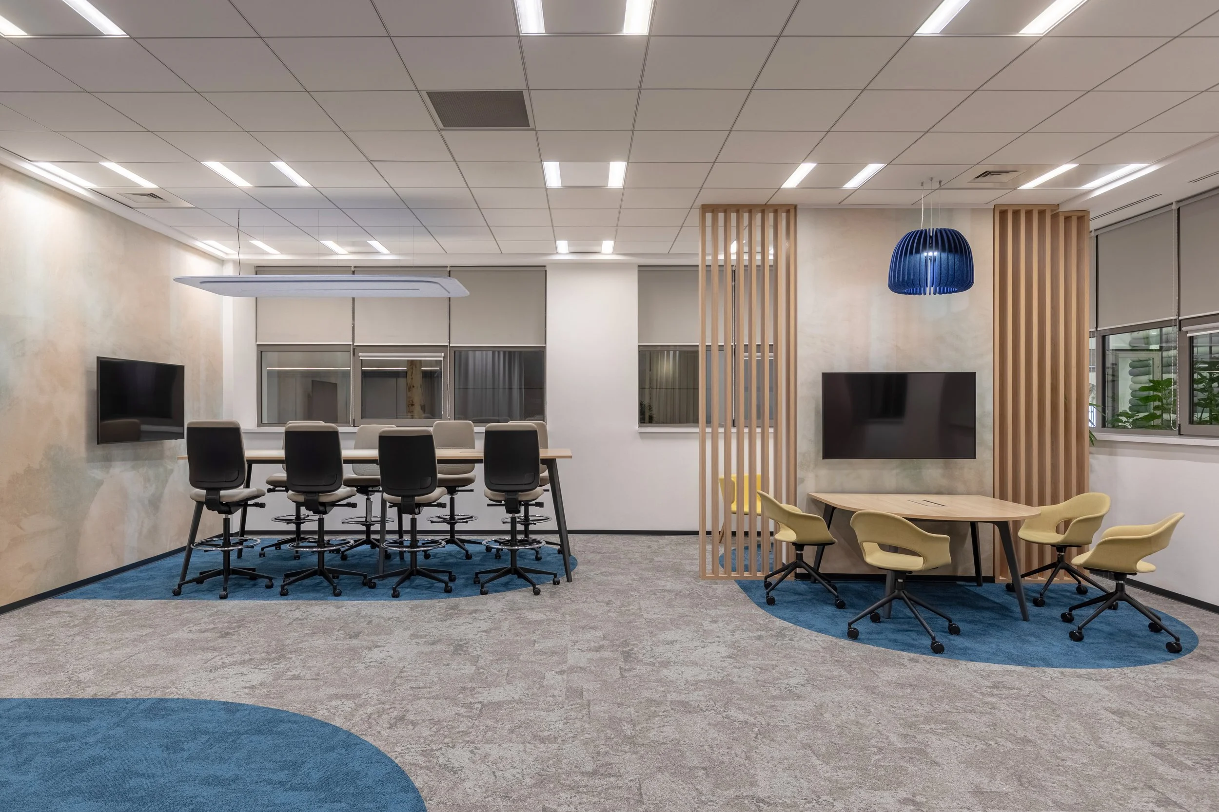 Modern office space with a high table and eight chairs on the left, and a small round meeting table with five yellow chairs on the right. A large flat-screen TV is mounted on the wall behind the meeting table, with a blue pendant light hanging above. Wooden room dividers partially separate the two areas, and windows with roller shades line the walls.