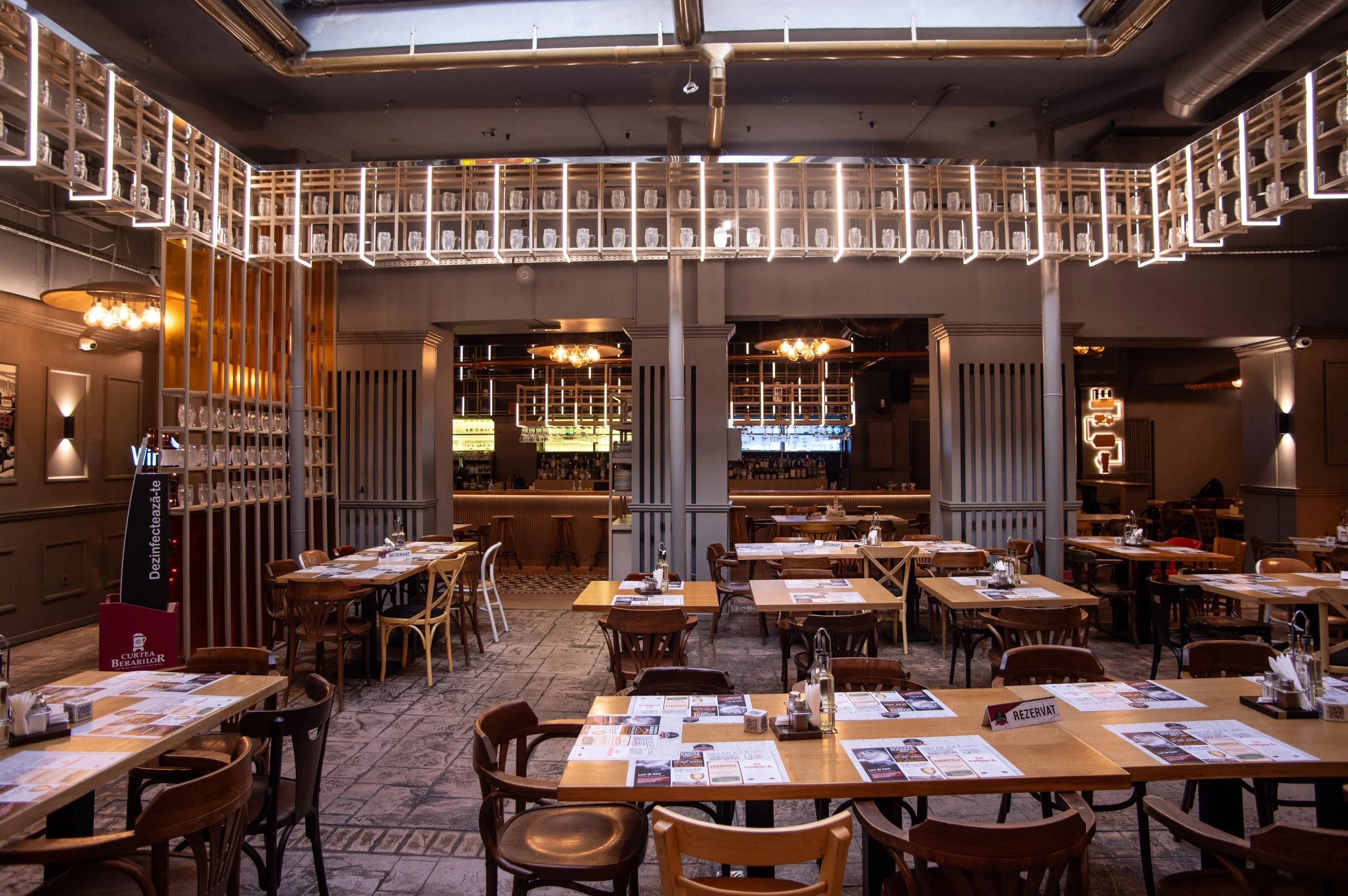 Interior of a modern restaurant with wooden tables and chairs, menu papers on the tables, decorative lighting fixtures, and a bar area in the background.
