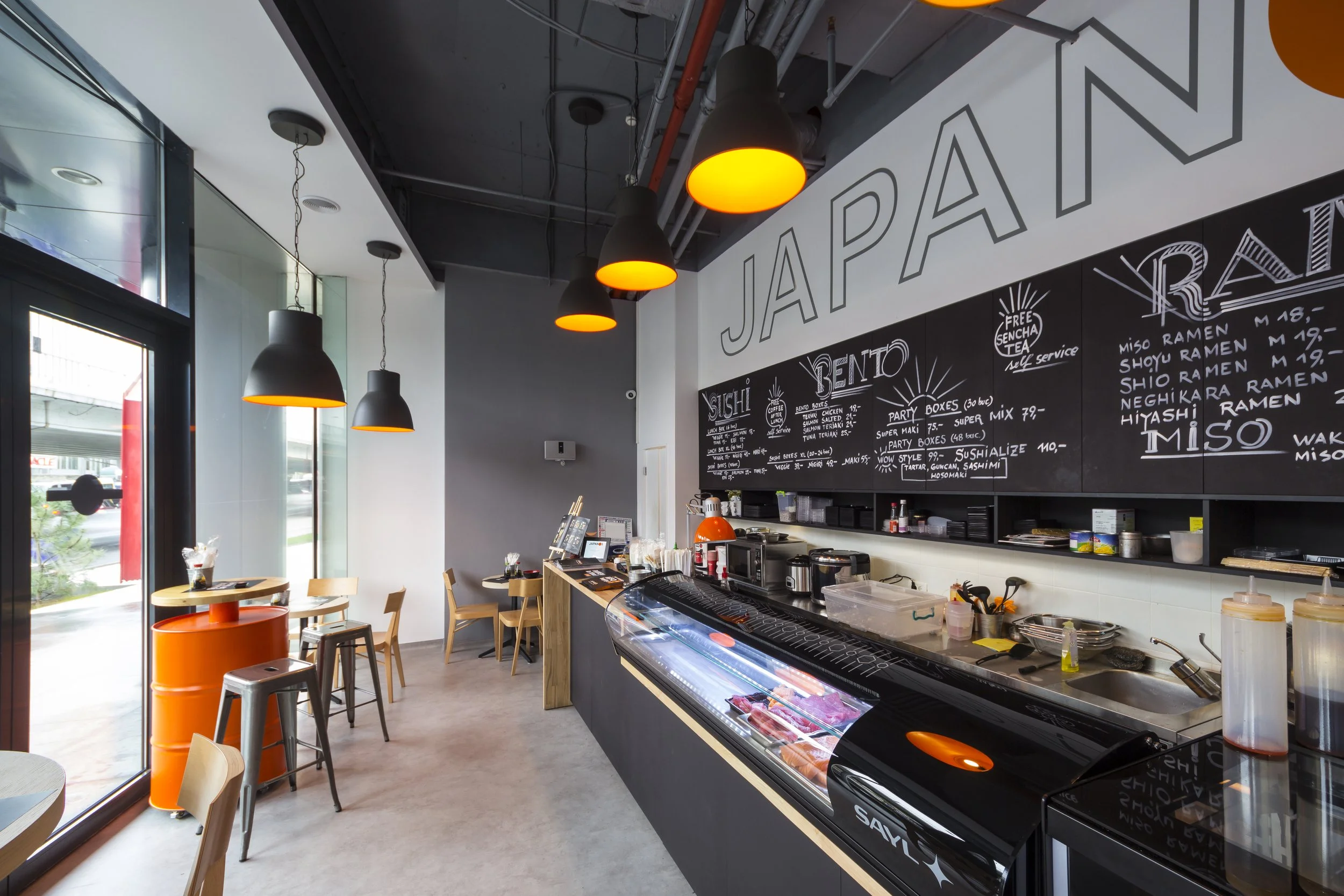 Interior of a modern Japanese restaurant with black chalkboard menu, gray walls, wooden tables, orange accents, and a counter displaying fresh meat.