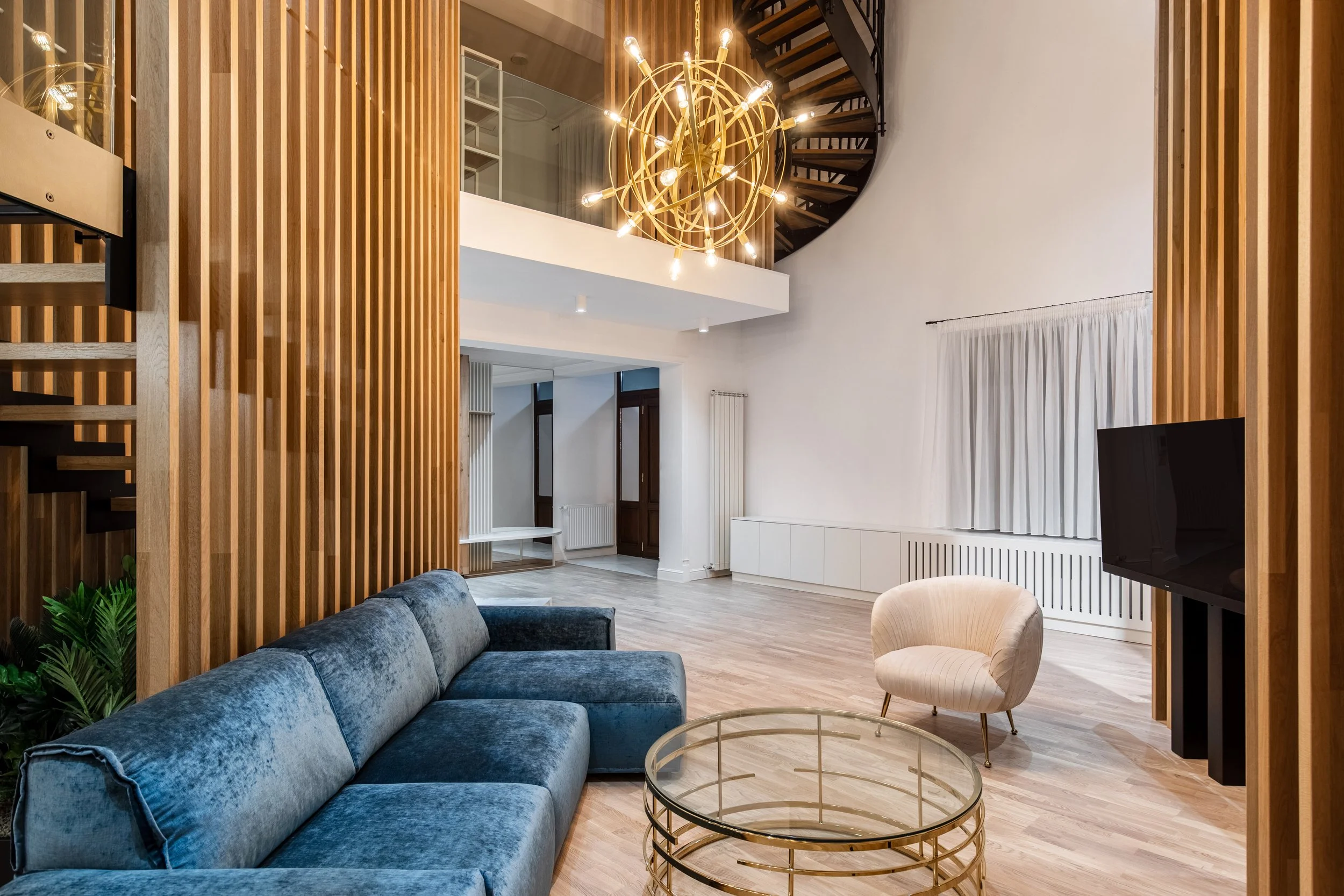 Modern living room with a blue velvet sofa, a round glass coffee table with gold frame, a white armchair, wooden slat wall, chandelier, spiral staircase, and large white curtain window.
