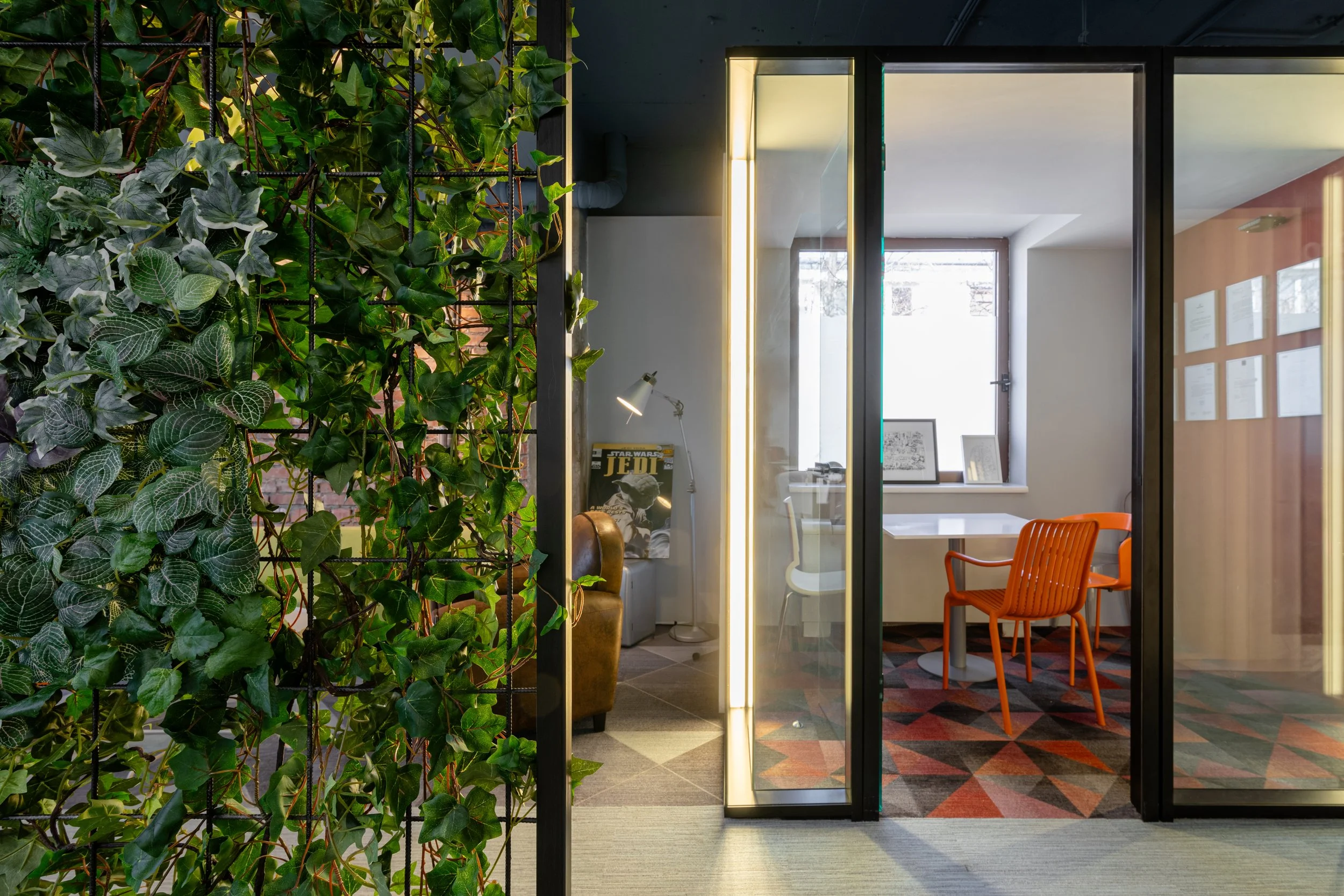 Indoor meeting room with orange chairs, framed photos, and a window on the back wall. Green houseplants on the left side and colorful geometric carpet on the floor.