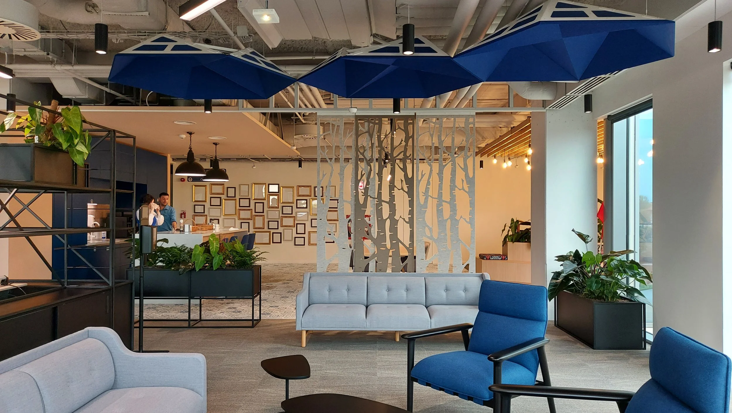Modern office lounge interior with blue umbrellas hanging from the ceiling, gray and blue chairs, indoor plants, and a decorative wooden screen dividing the spaces.