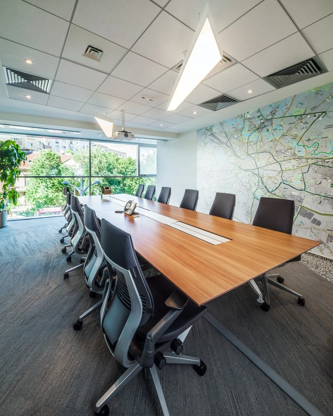 A modern conference room with a long wooden table, black ergonomic chairs, large window with city view, map on the wall, ceiling lights, and a projector.