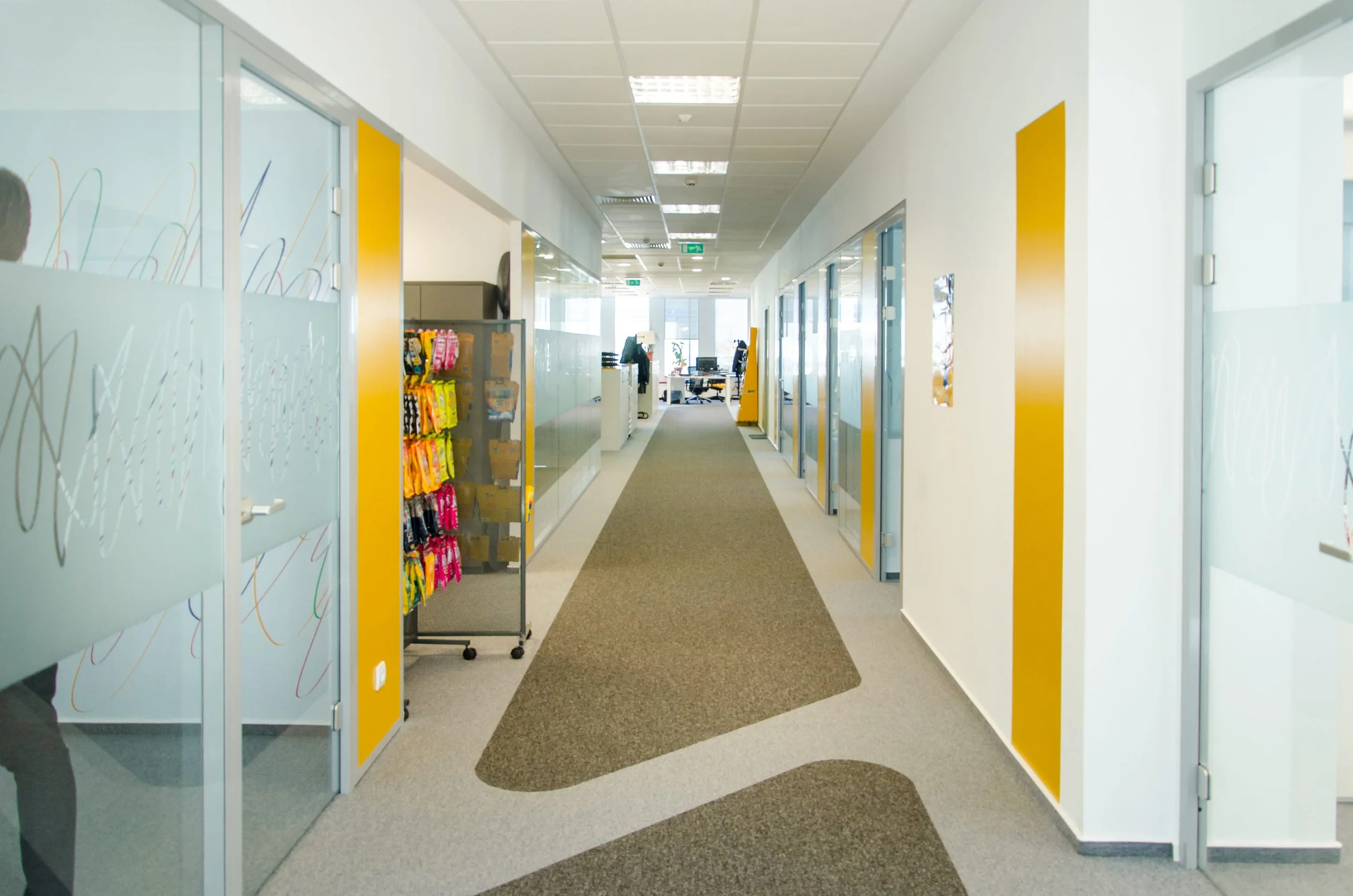 Office corridor with glass-walled rooms on one side and plain walls on the other, yellow accent panels, a gray and brown patterned carpet, and a person working at a desk in the distance.