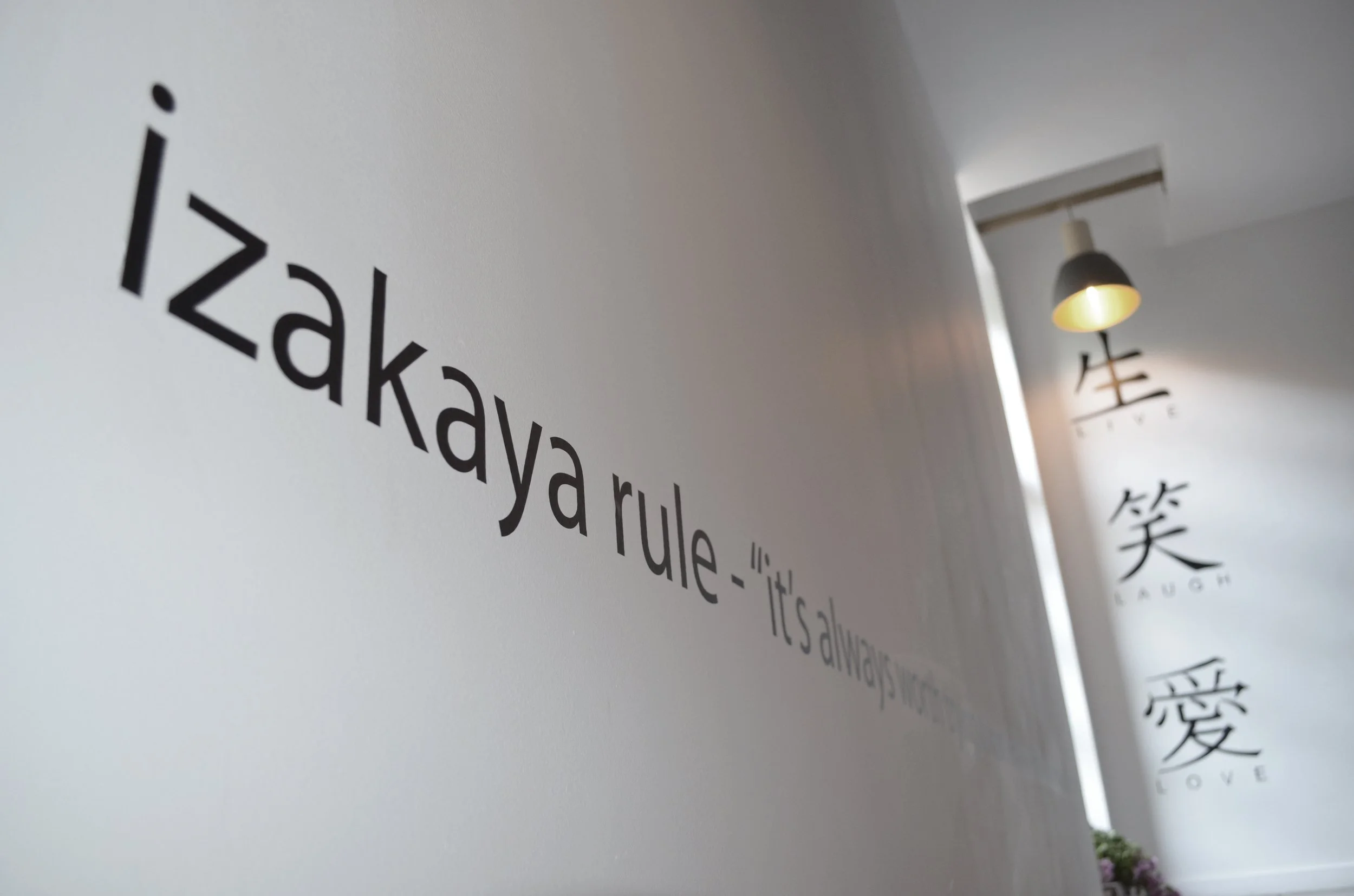 Wall with the phrase 'izakaya rule - it's always love' and hung artwork displaying the word 'LOVE' along with Japanese characters for love, beneath a hanging lamp.