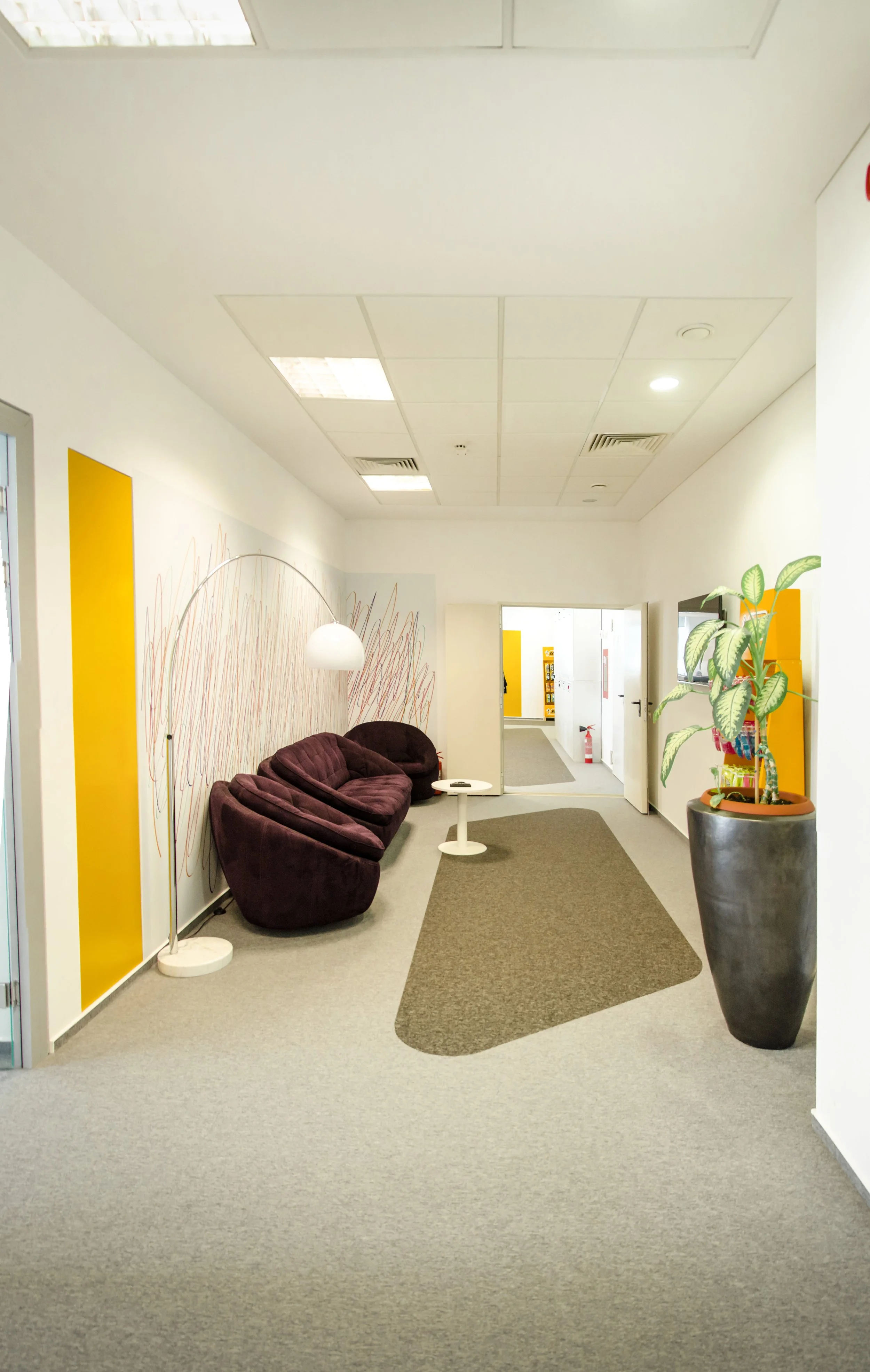 Modern office waiting area with purple couch, white side table, large potted plant, yellow accent wall, and artwork on white walls.