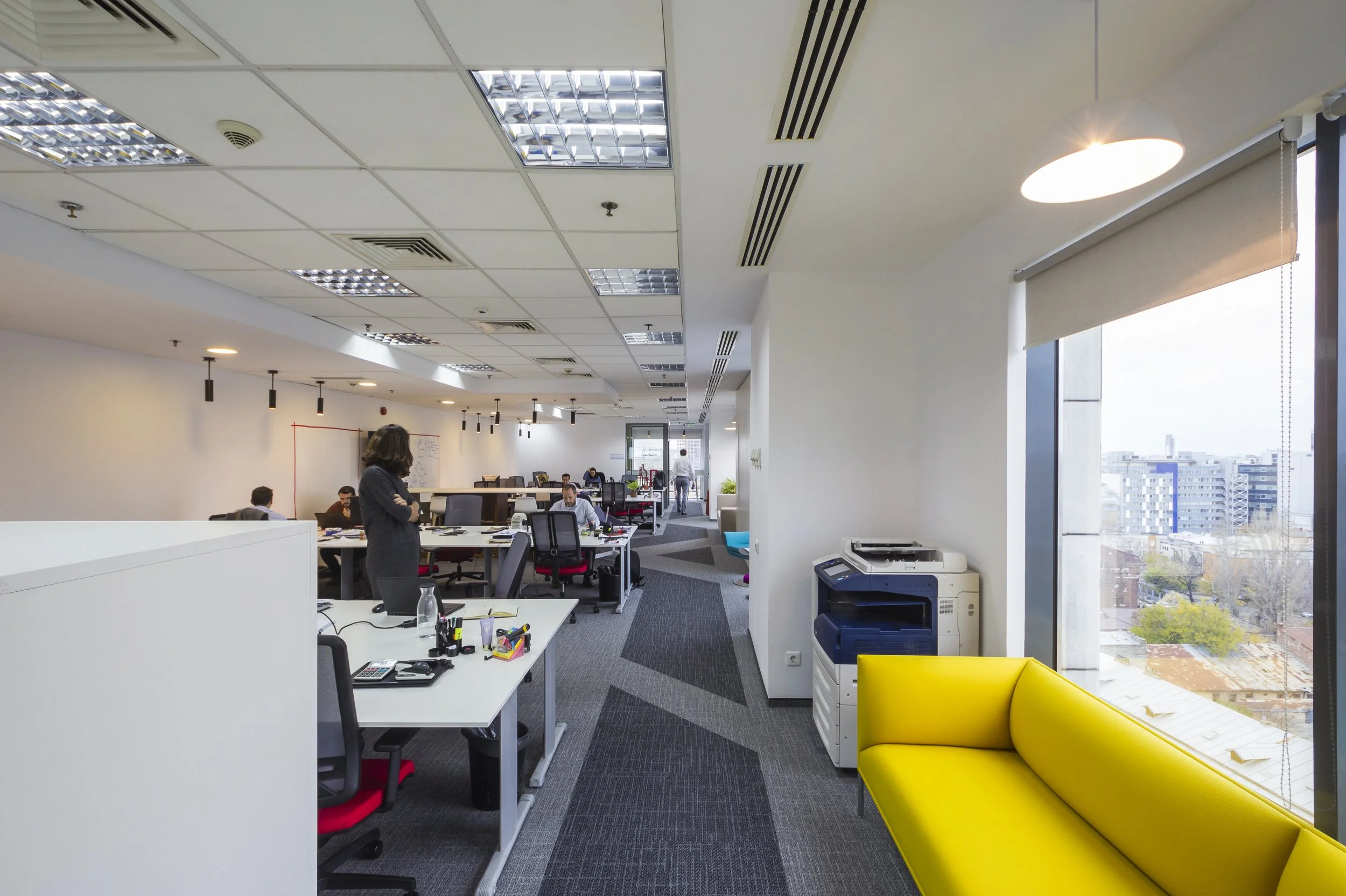 Modern open-plan office with desks, chairs, a yellow sofa near large windows, and people working at their desks.