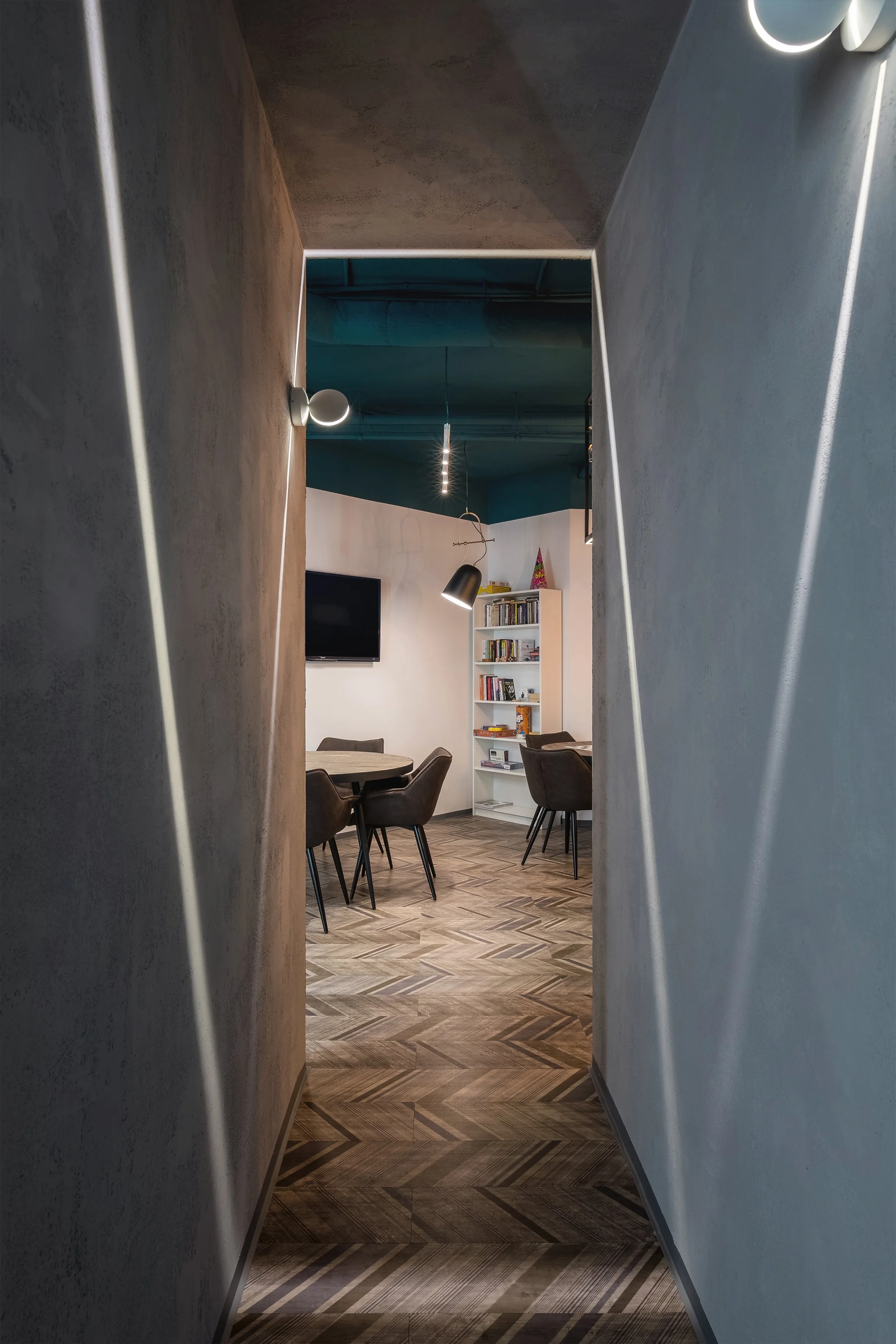 A hallway leading to a room with a round table, six chairs, a bookshelf, a wall-mounted television, and contemporary lighting fixtures, with a wood-patterned floor.