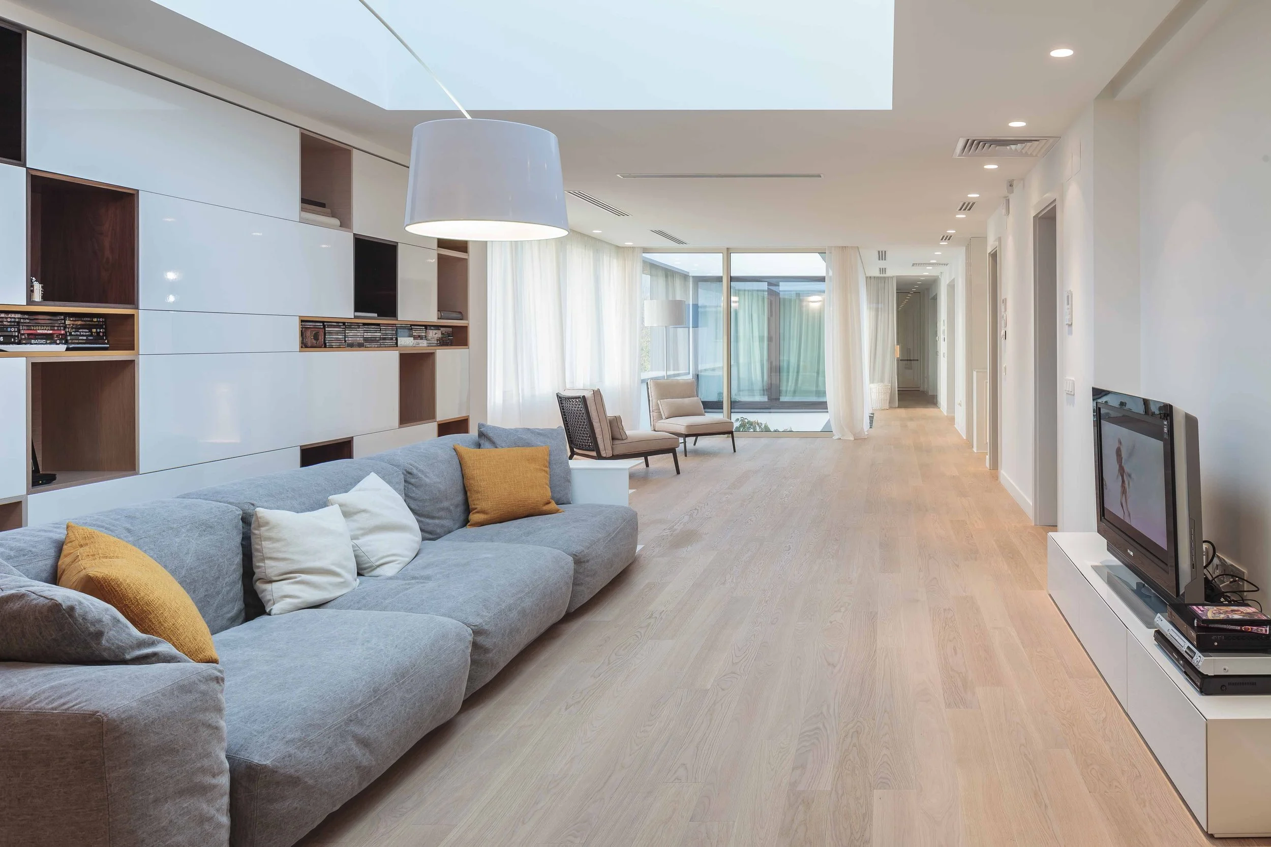 Modern living room with gray sofa, yellow and white pillows, wooden flooring, built-in wall shelves, a large pendant light, and a sitting area near large windows with white curtains.